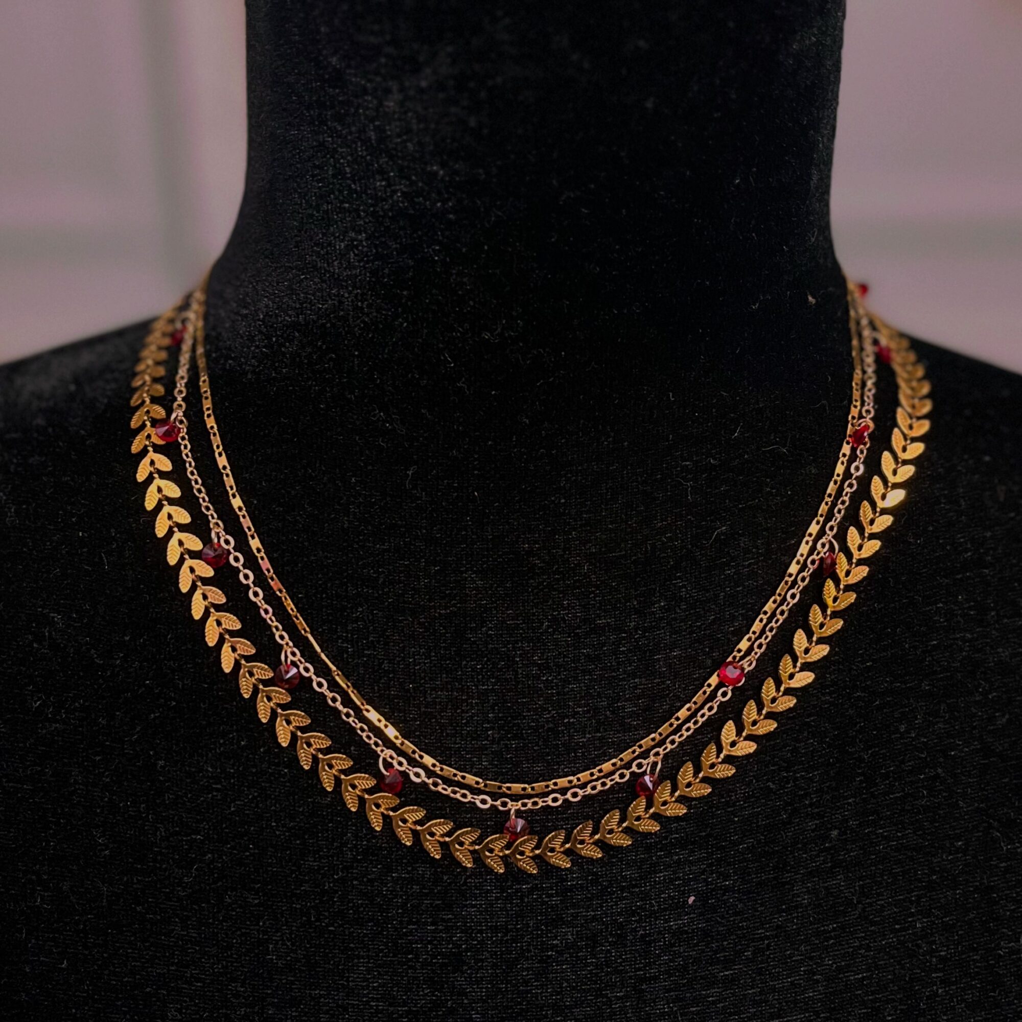 Three layered gold necklaces with leaf and chain designs displayed on a black bust, with a blurred background.