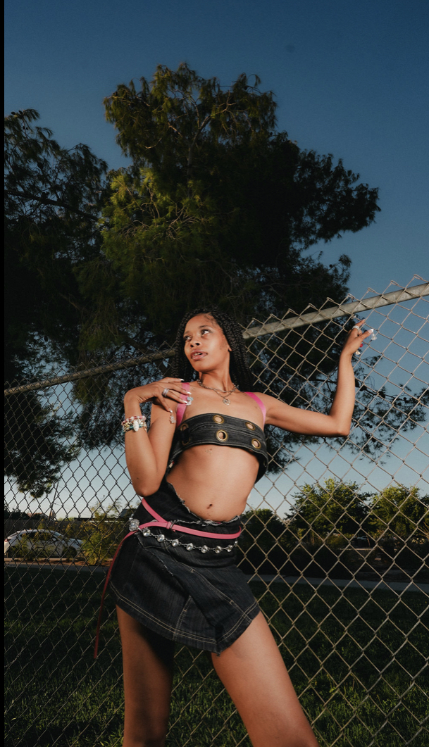 Young woman standing outdoors near a chain-link fence, with trees and a blue sky in the background.