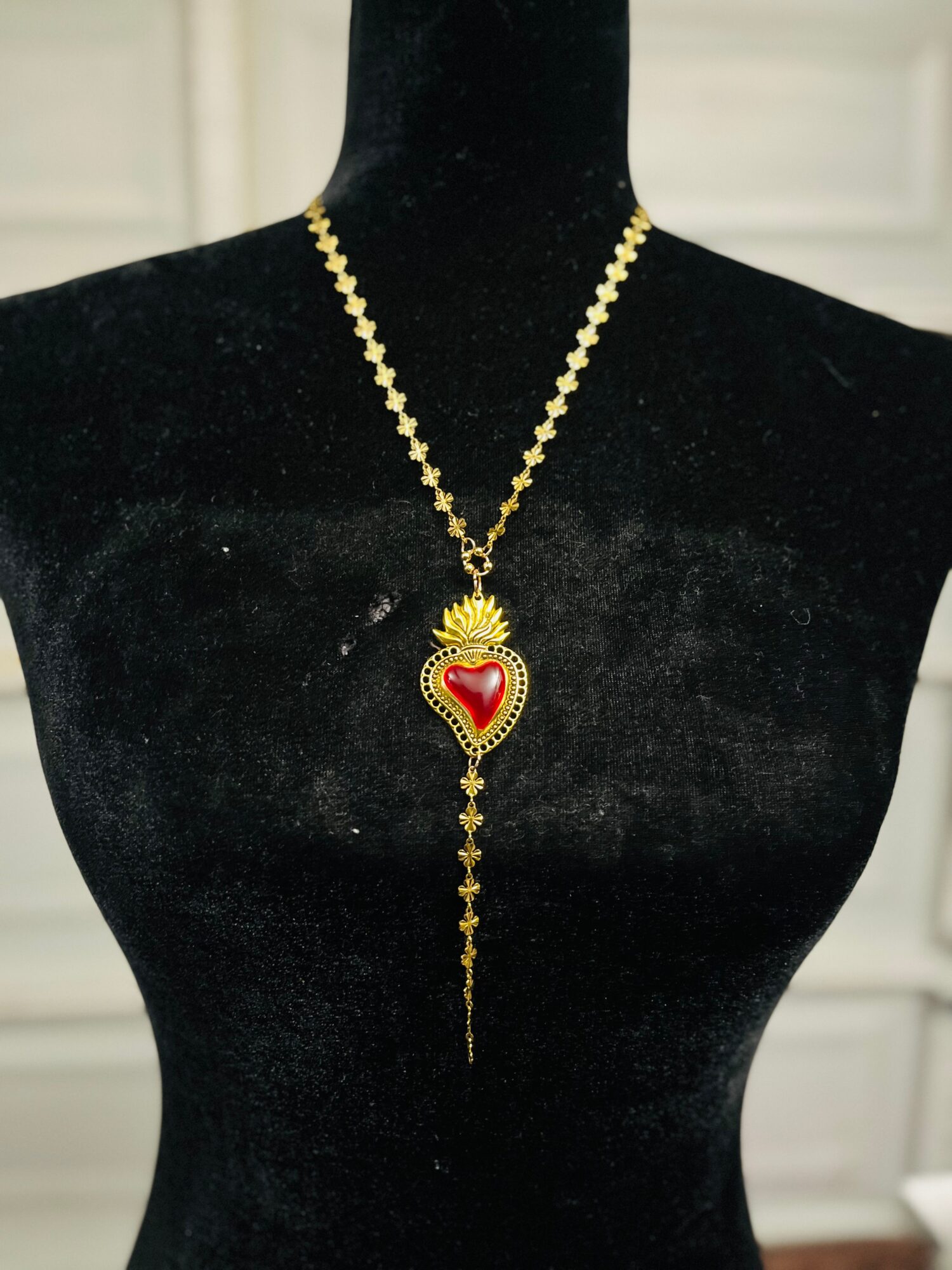 Gold necklace with a heart-shaped pendant featuring a red center and a crown design, hanging from a black display bust.
