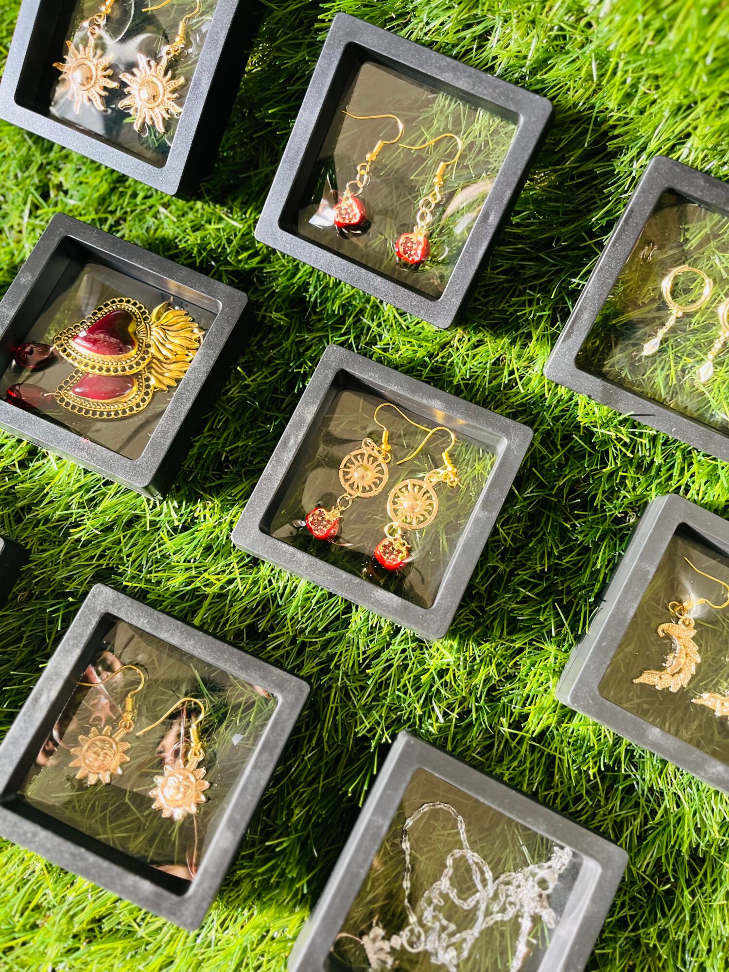 Jewelry displayed in black boxes on green grass, including necklaces, earrings, and rings with gold and red accents.
