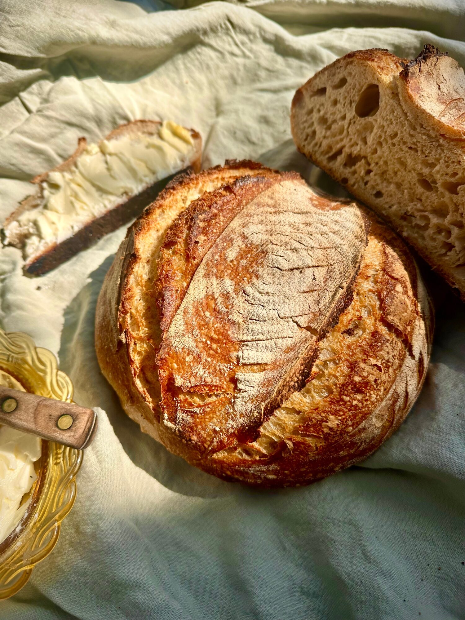Round loaf of bread with a crusty exterior and scored top, partially sliced to reveal soft interior, on fabric surface.