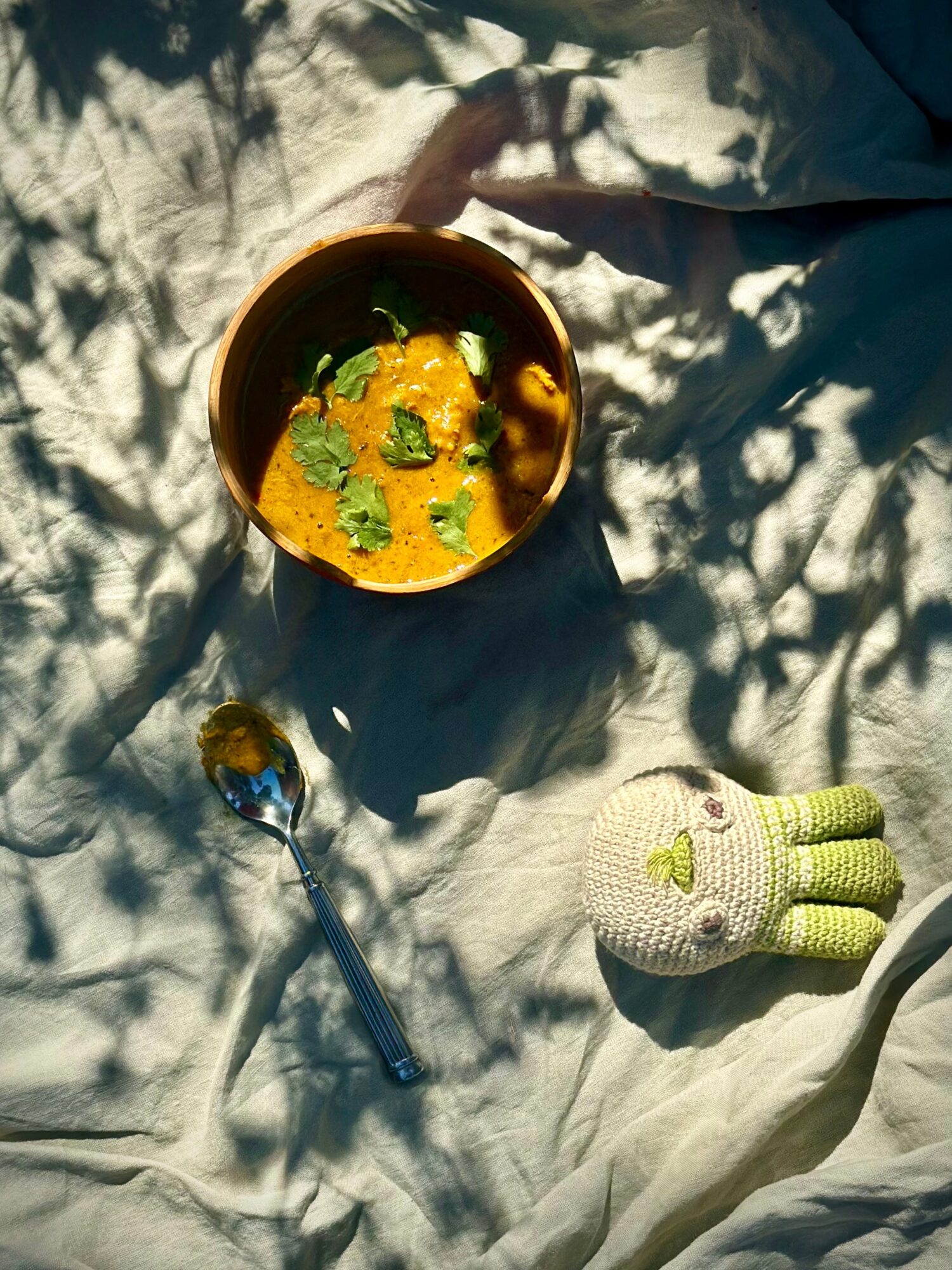 Bowl of yellow curry with green herbs, spoon, and a stuffed toy on a light-colored fabric surface with shadows of leaves.