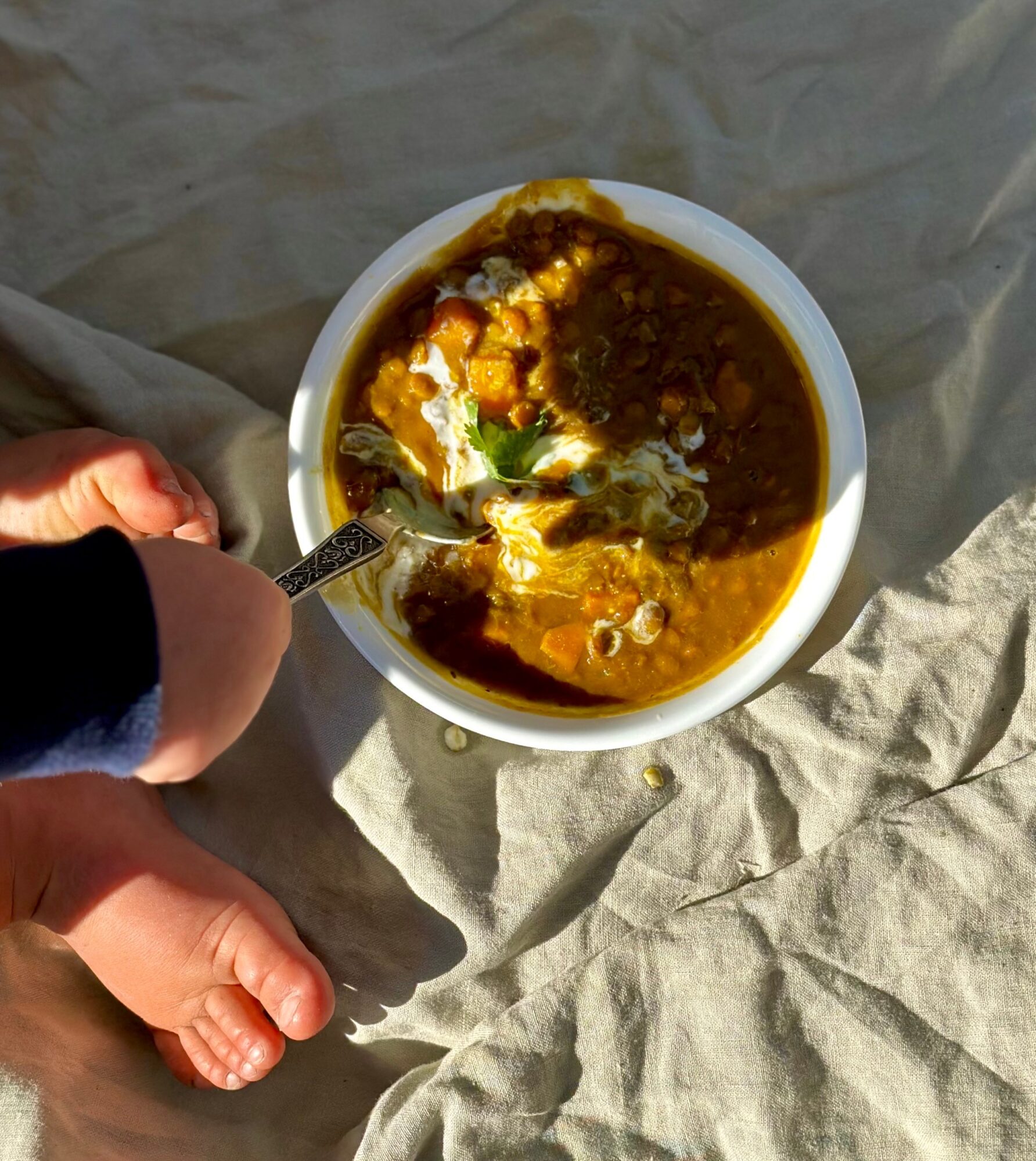 Person holding a spoon over a bowl of soup with vegetables, on crumpled paper surface.