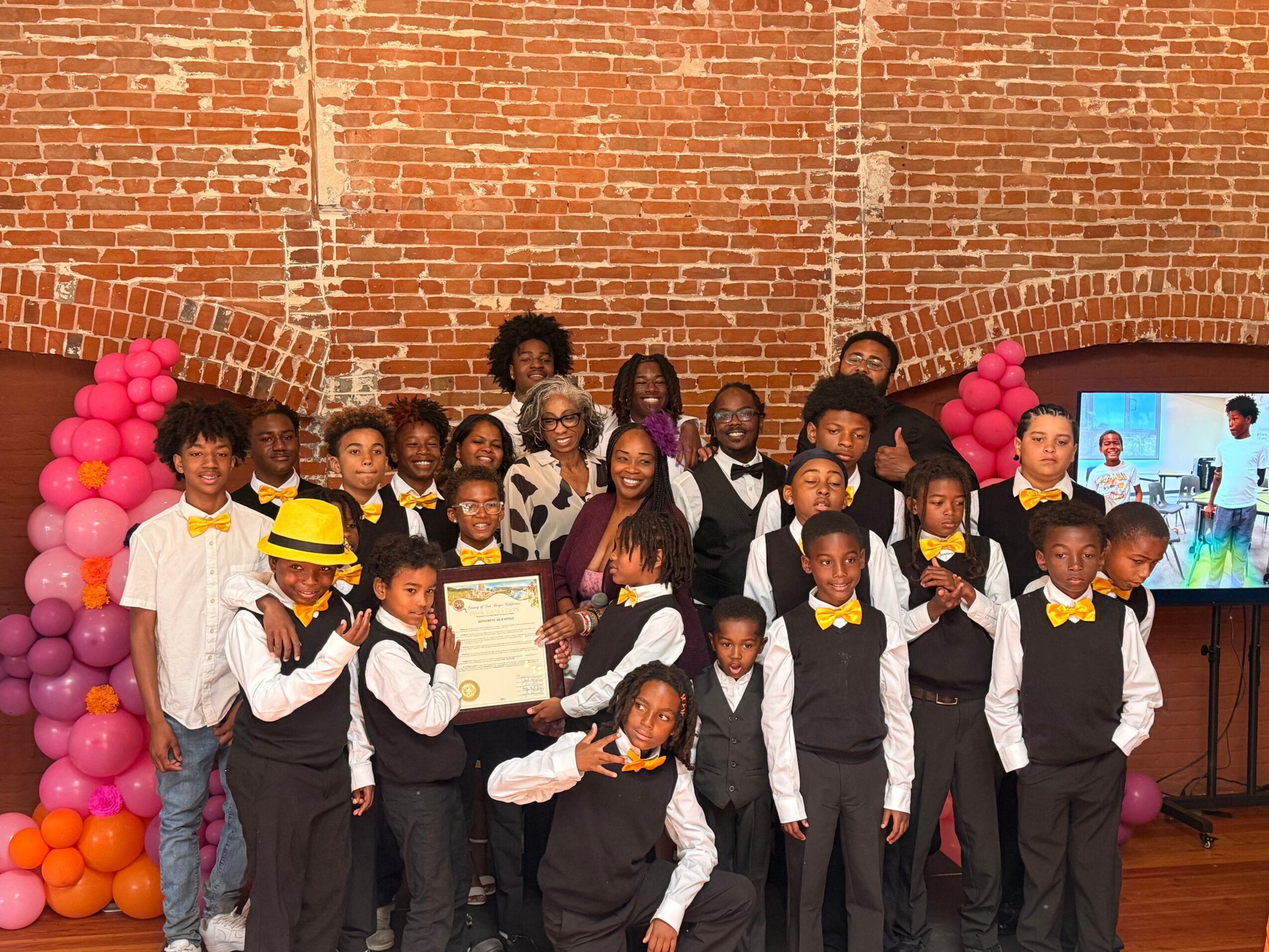 Group of children and adults in formal attire standing in front of a brick wall, with pink balloon decorations and a screen displaying a presentation.