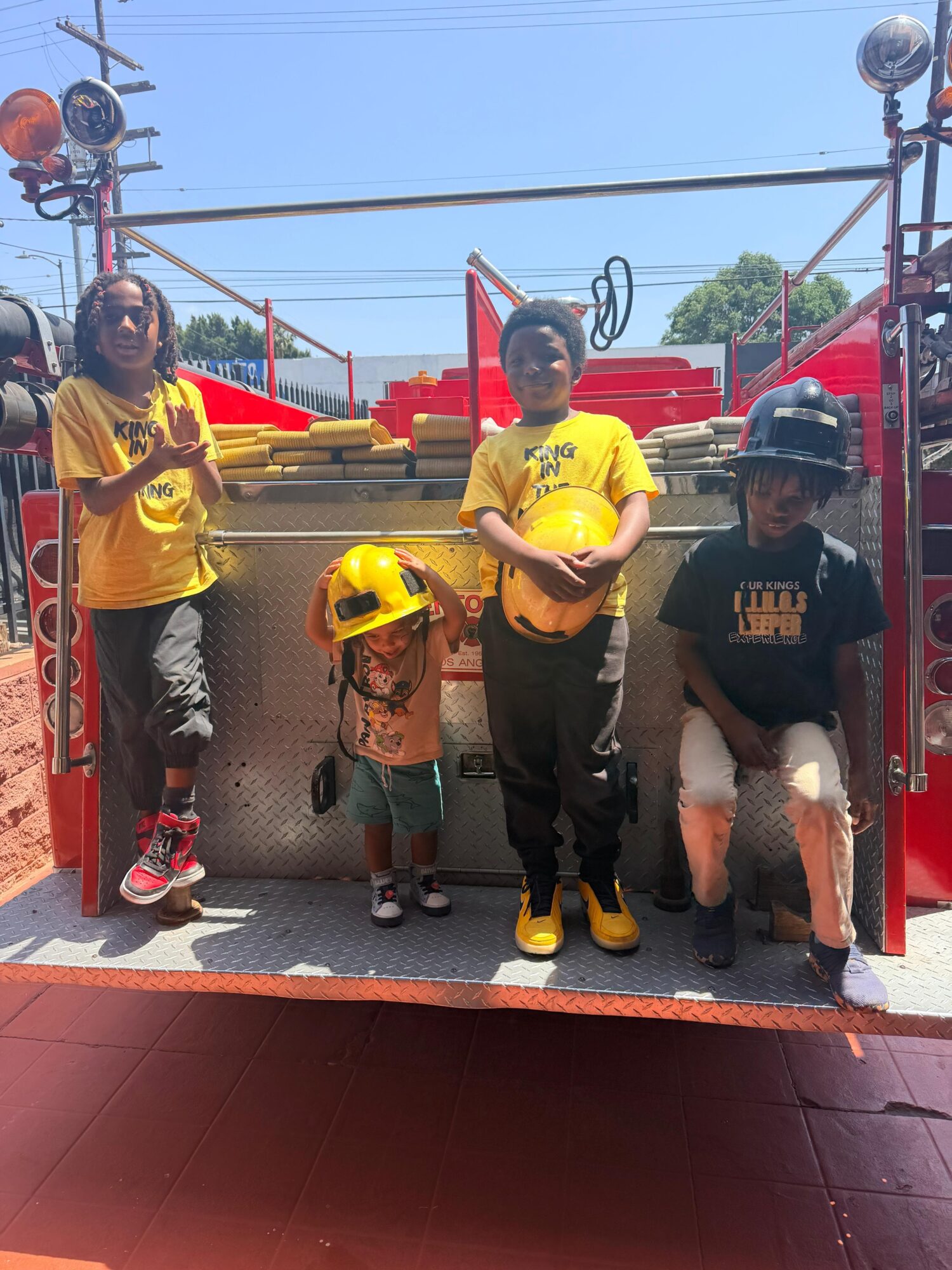 Four children stand on a fire truck, one holding a yellow helmet, outdoors on a sunny day.