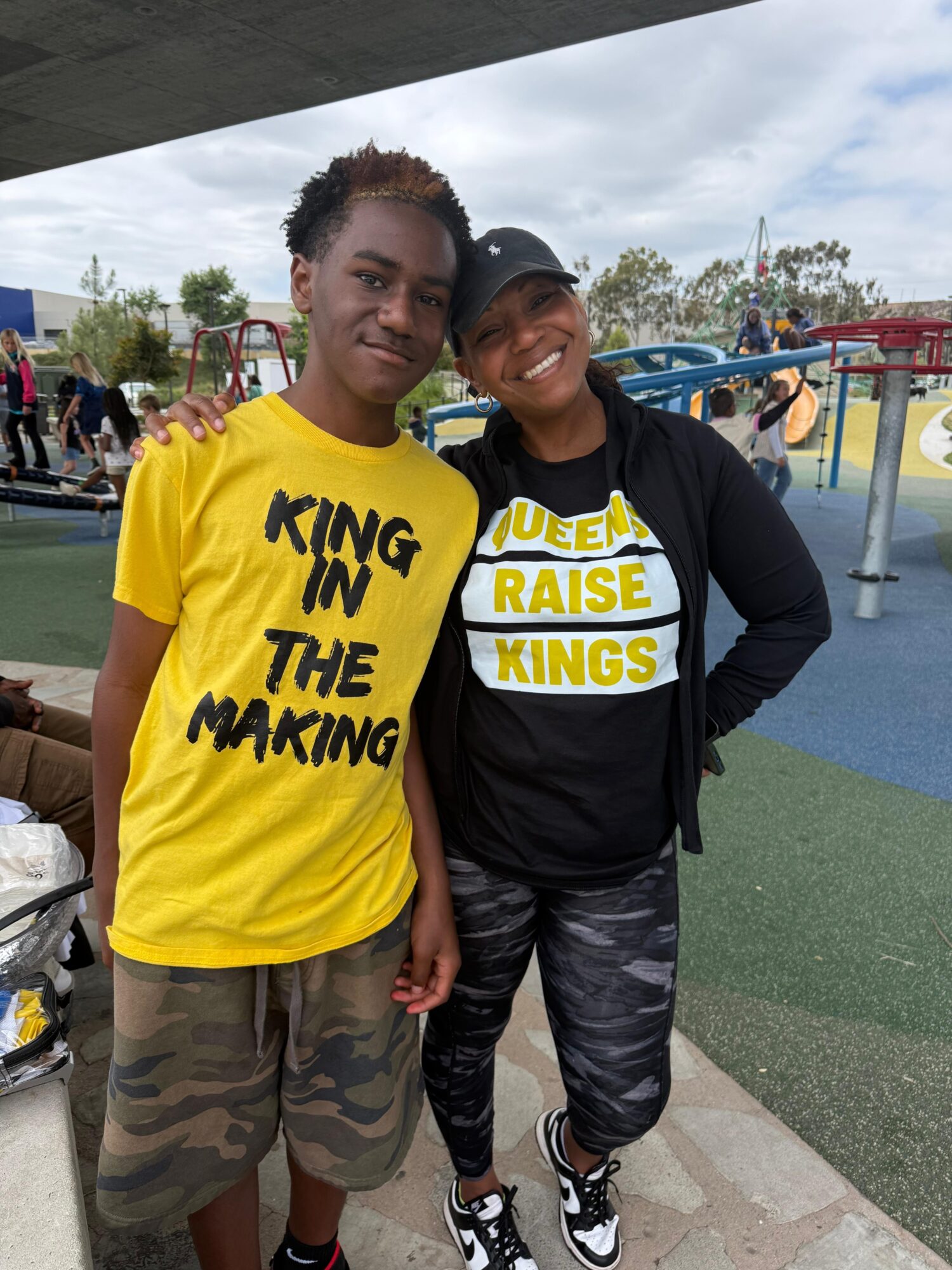 Two people smiling at an outdoor playground, one wearing a yellow shirt and the other in black, with colorful play structures in background.