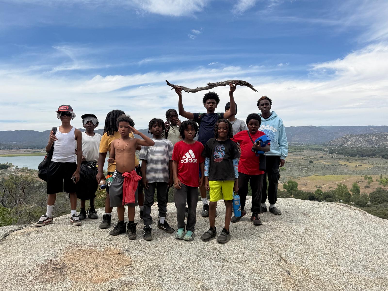Group of children and a person holding a branch on a rocky hilltop with a landscape and blue sky in background.