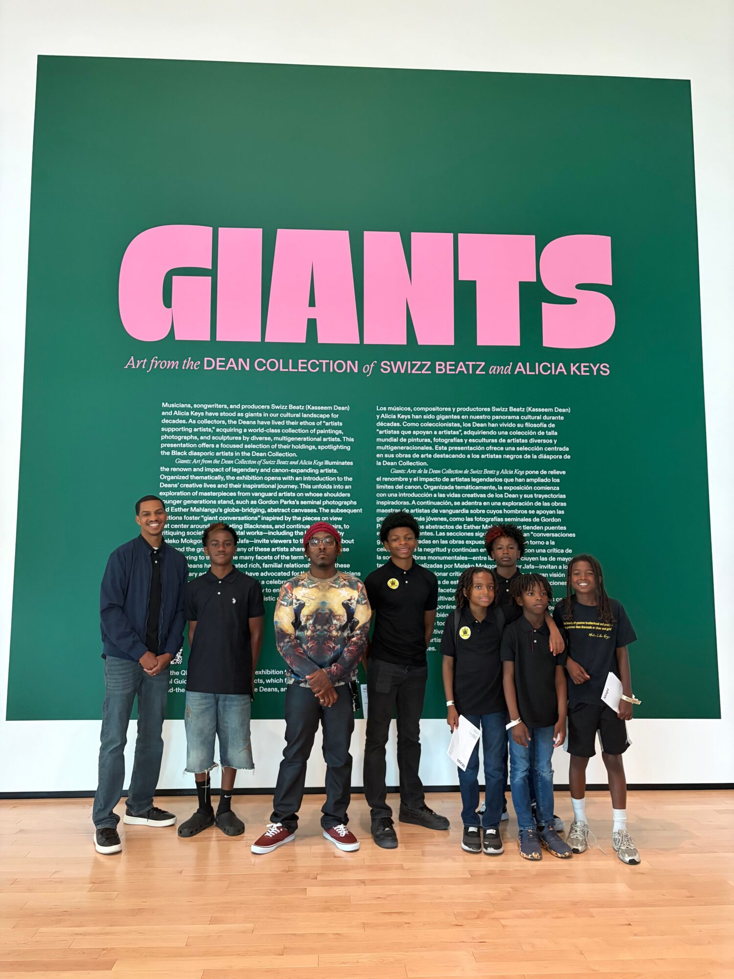 Group of six people standing in front of a large green display with pink and white text, on a wooden floor.