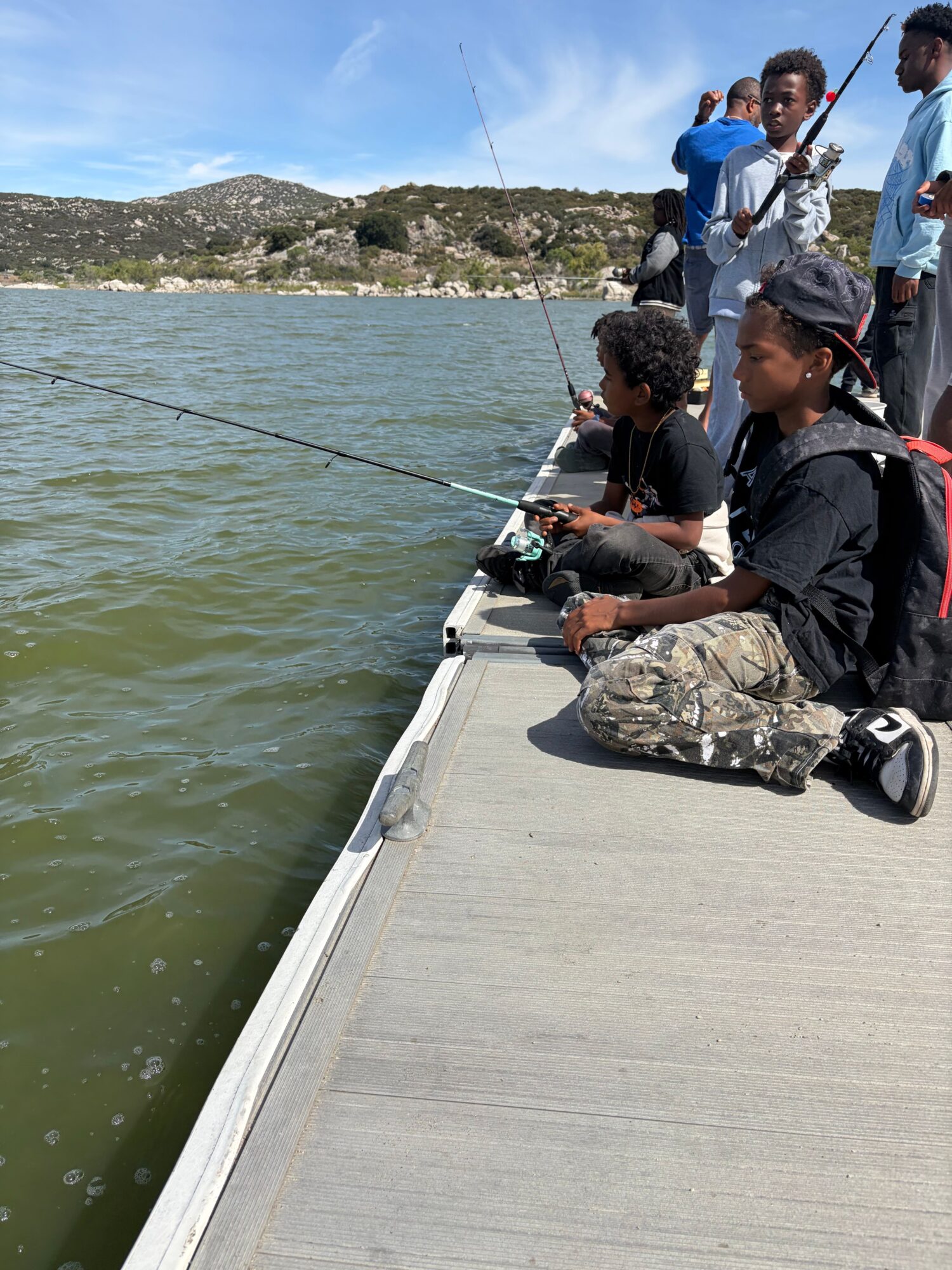 Group of people fishing from a dock over water, some holding fishing rods, with a landscape background.