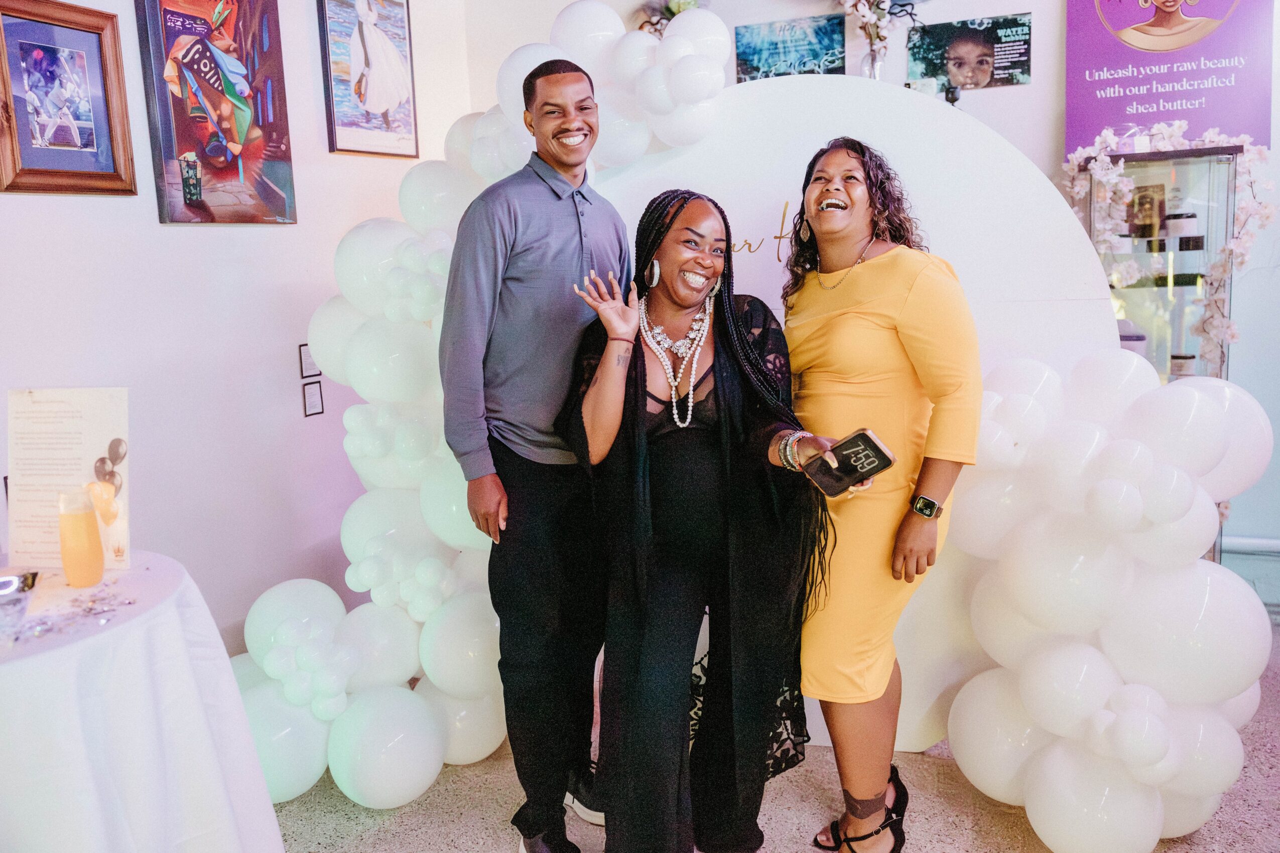 Three people smiling and waving in front of a cloud-shaped balloon backdrop at an indoor event, with colorful artwork on the wall.