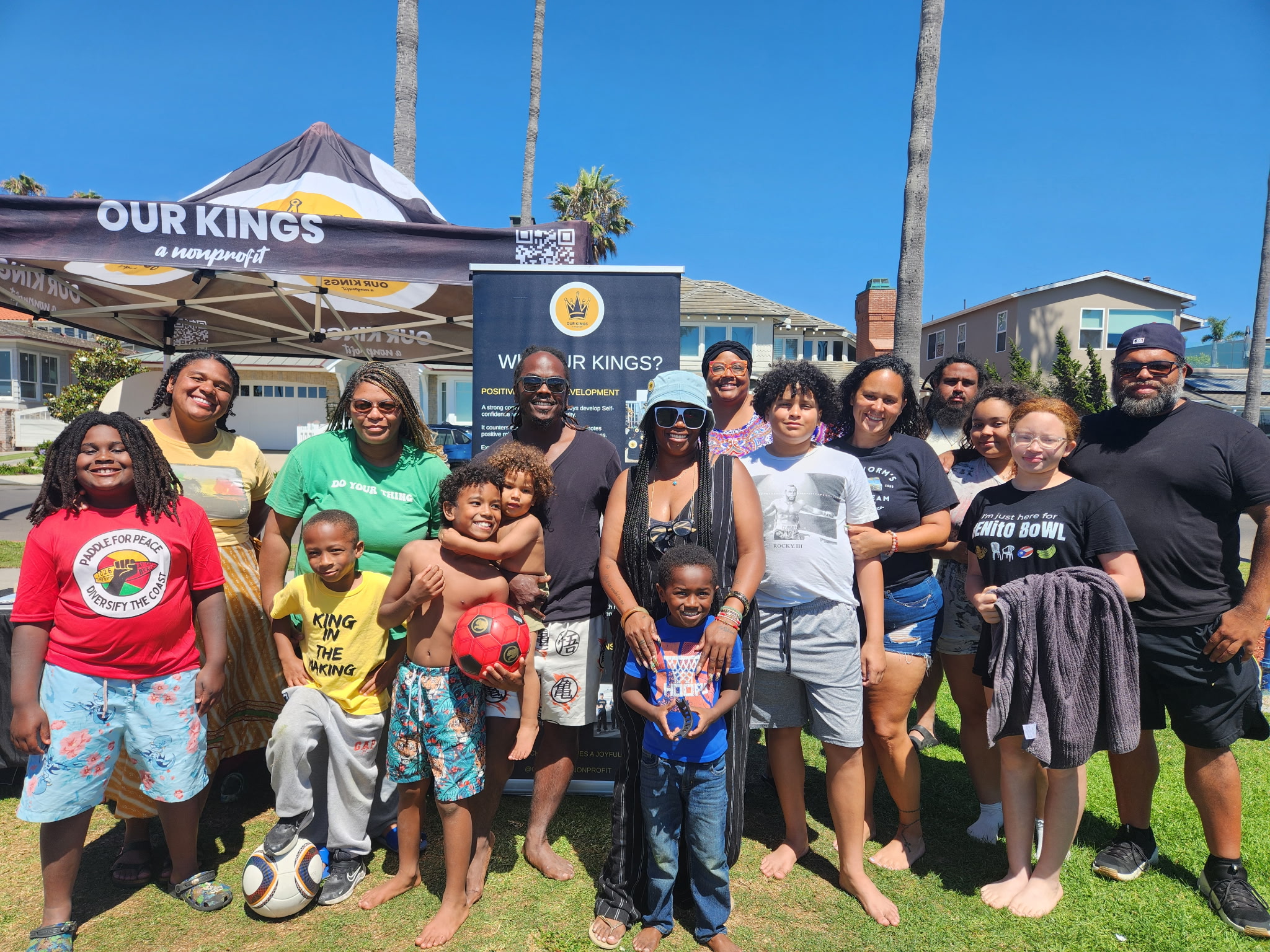 Group of people, including children, standing outdoors in front of a tent and a sign, smiling for a photo.