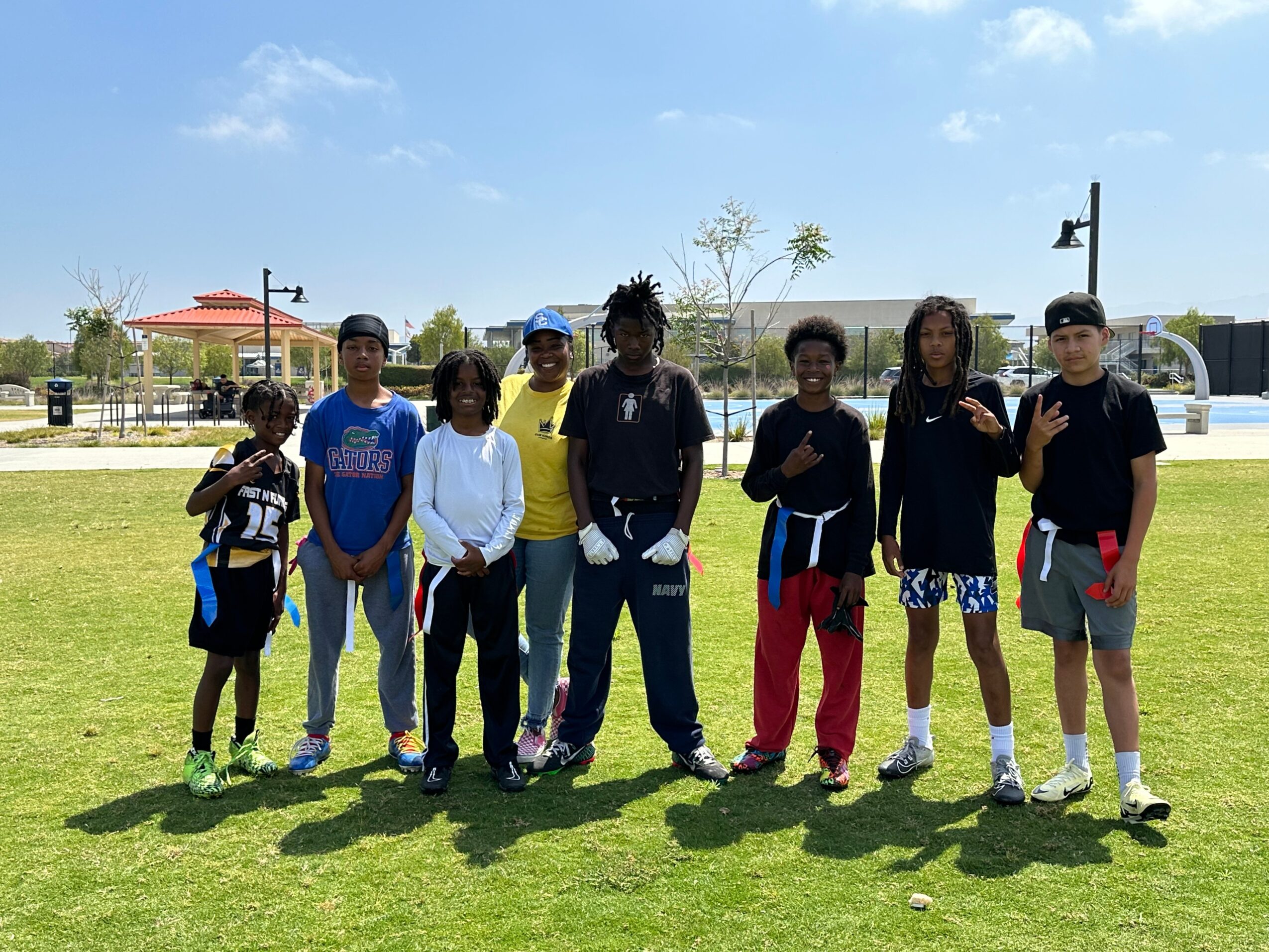 Group of nine children standing on grass in a park, with trees, buildings, and a blue sky in the background.