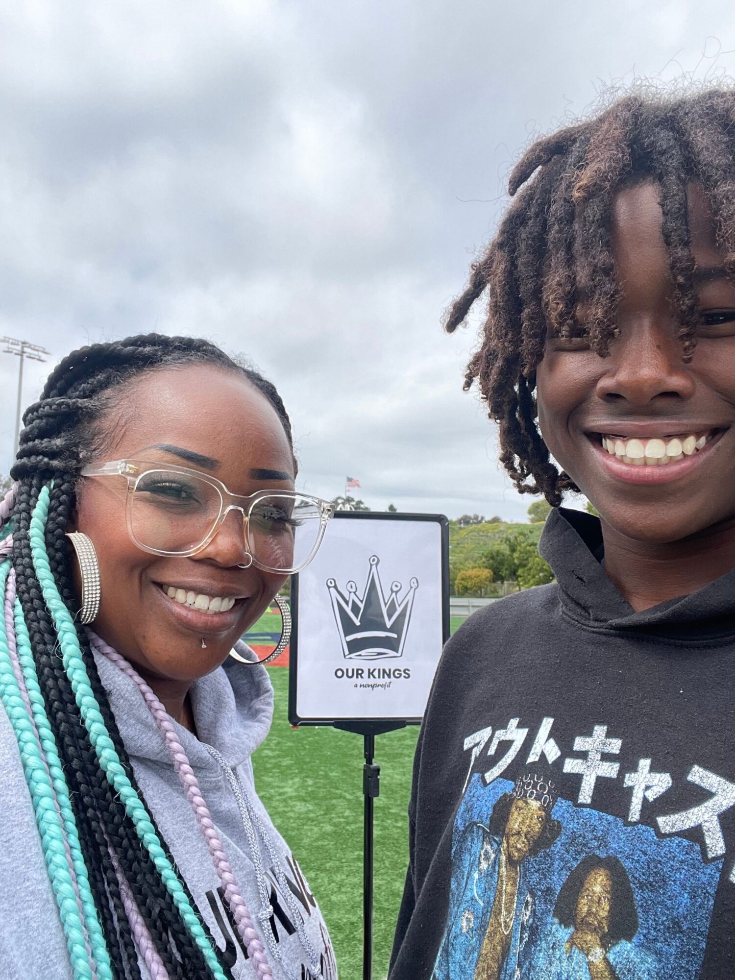 Two smiling people outdoors, one with glasses and braided hair, the other with dreadlocks, standing near a sign on a grassy field.