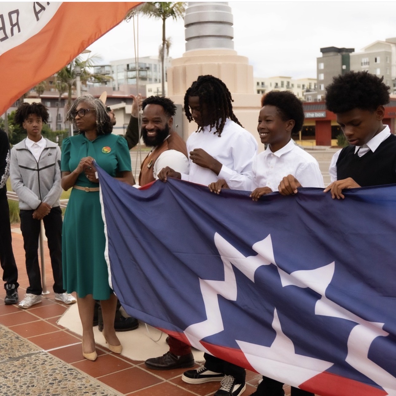 Group of people holding a flag outdoors, with buildings and trees in the background.