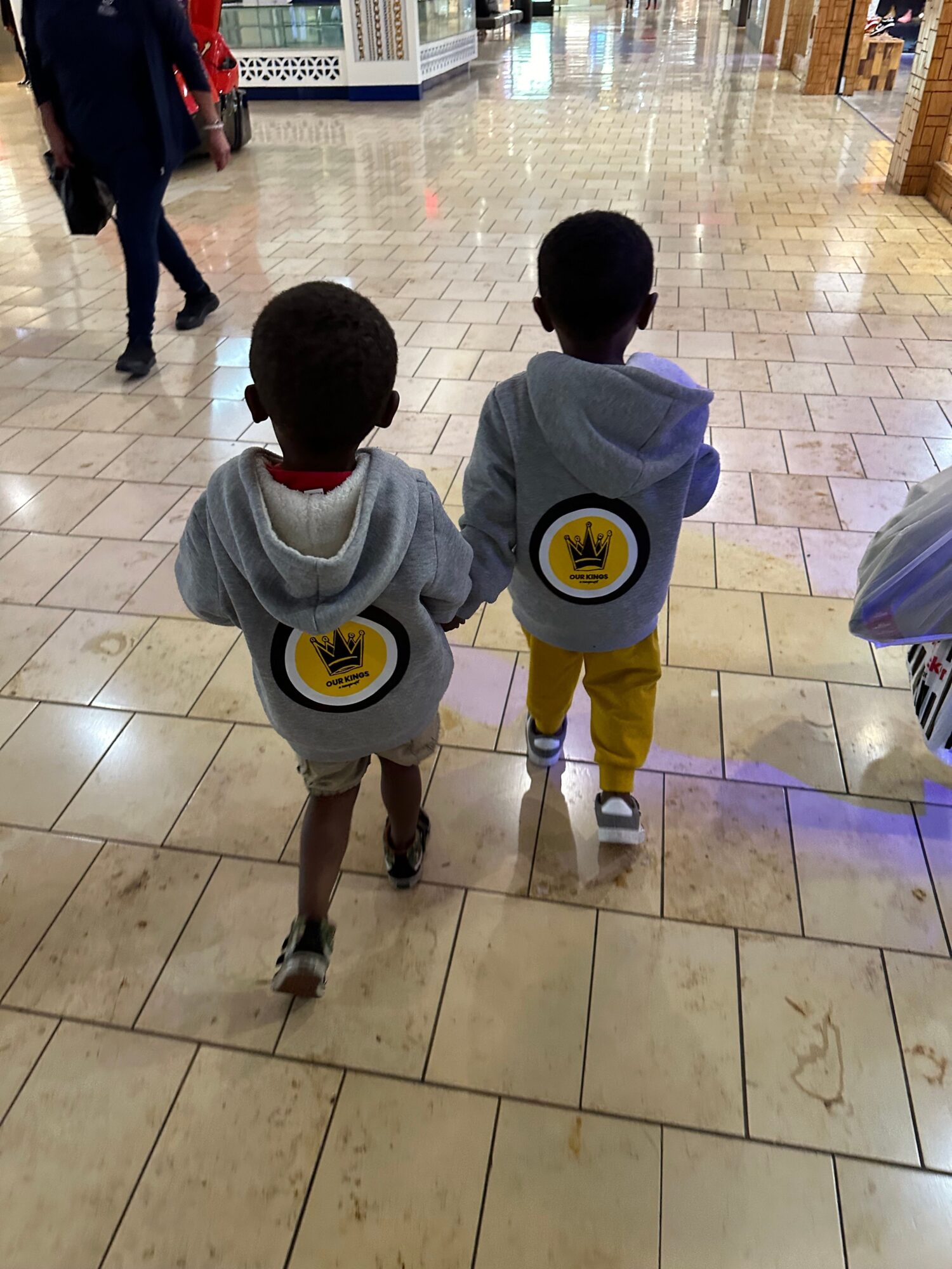 Two children walking on tiled floor, one with a backpack, in a shopping mall or public space.