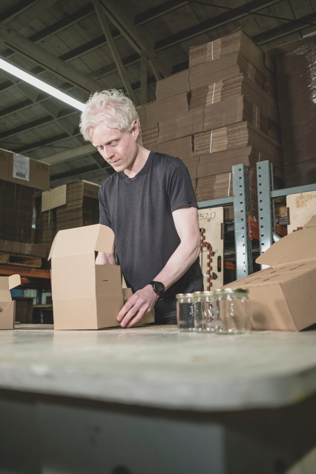 Person with curly gray hair in black shirt packing boxes in warehouse, shelves and stacked boxes in background.