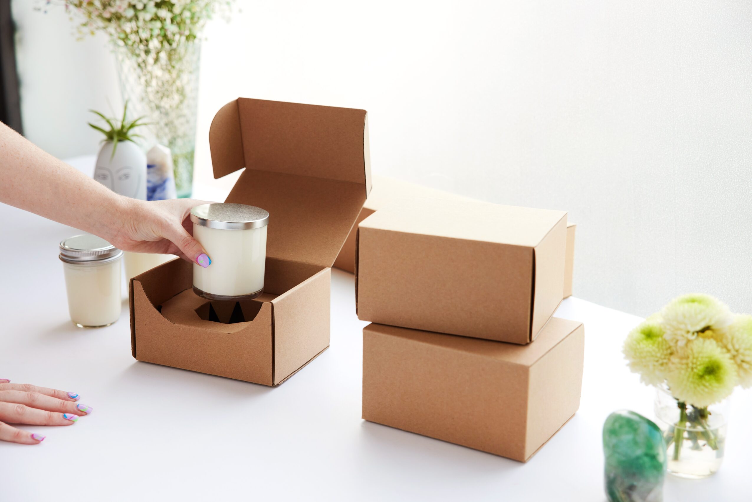 Person placing a jar into a cardboard box, with two more boxes nearby and flowers in the background.