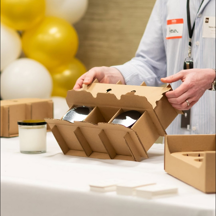 Person opening a cardboard box with three glass jars inside, balloons in the background, on a table with a candle and other boxes.