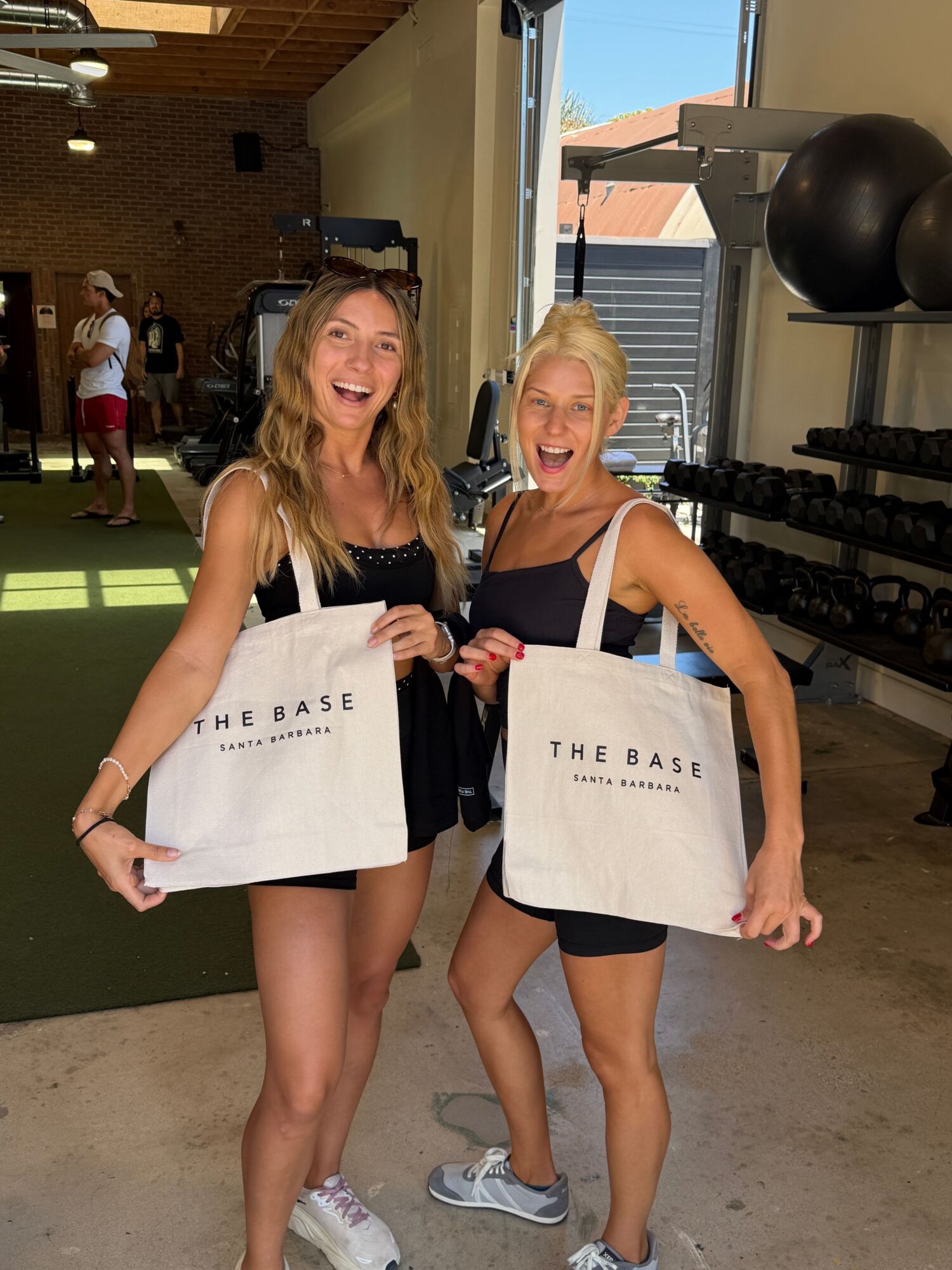 Two women holding shopping bags in a gym, smiling, with gym equipment in the background.