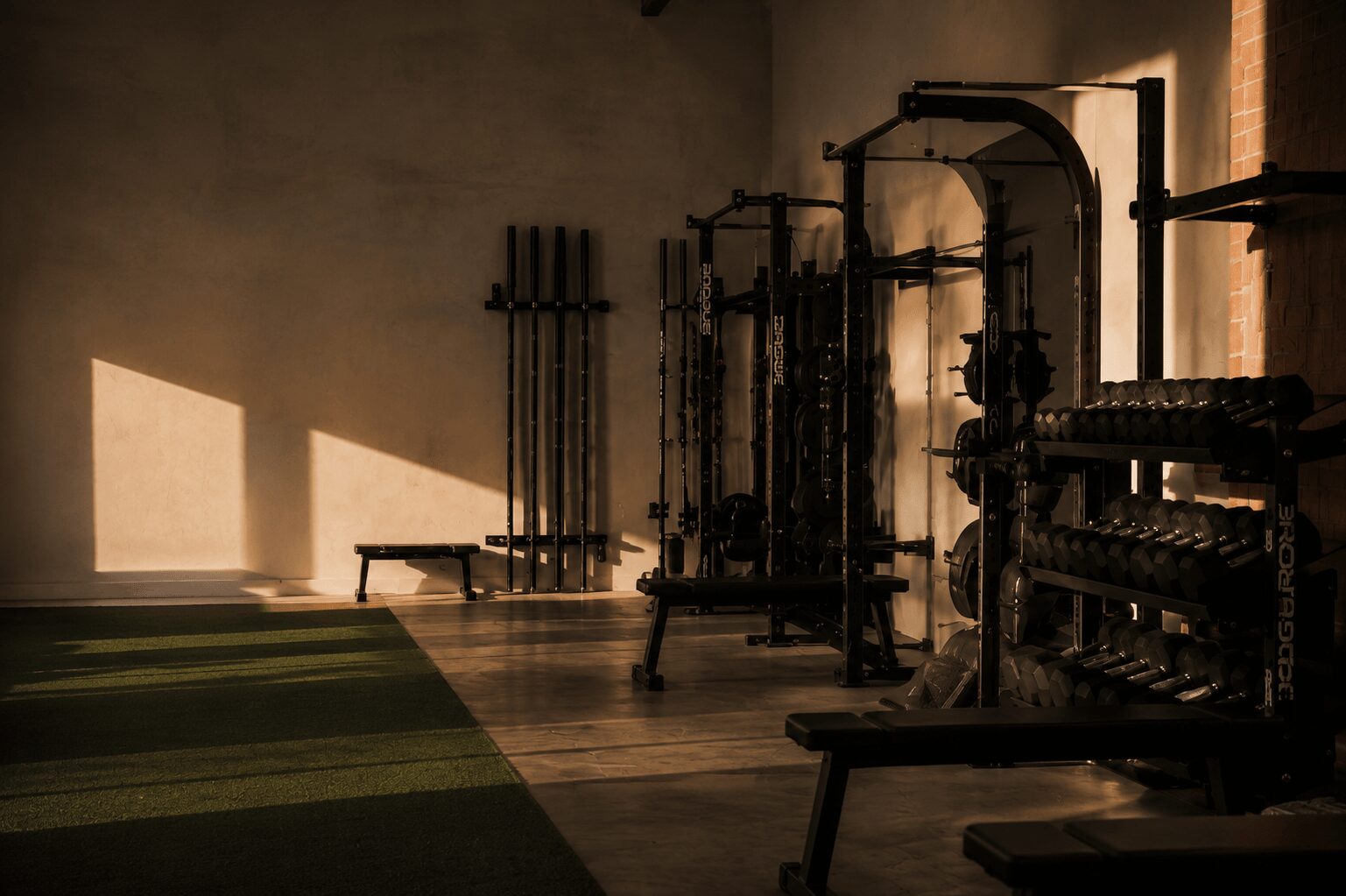 Gym equipment in a dimly lit room with sunlight streaming through windows, casting shadows on the floor.