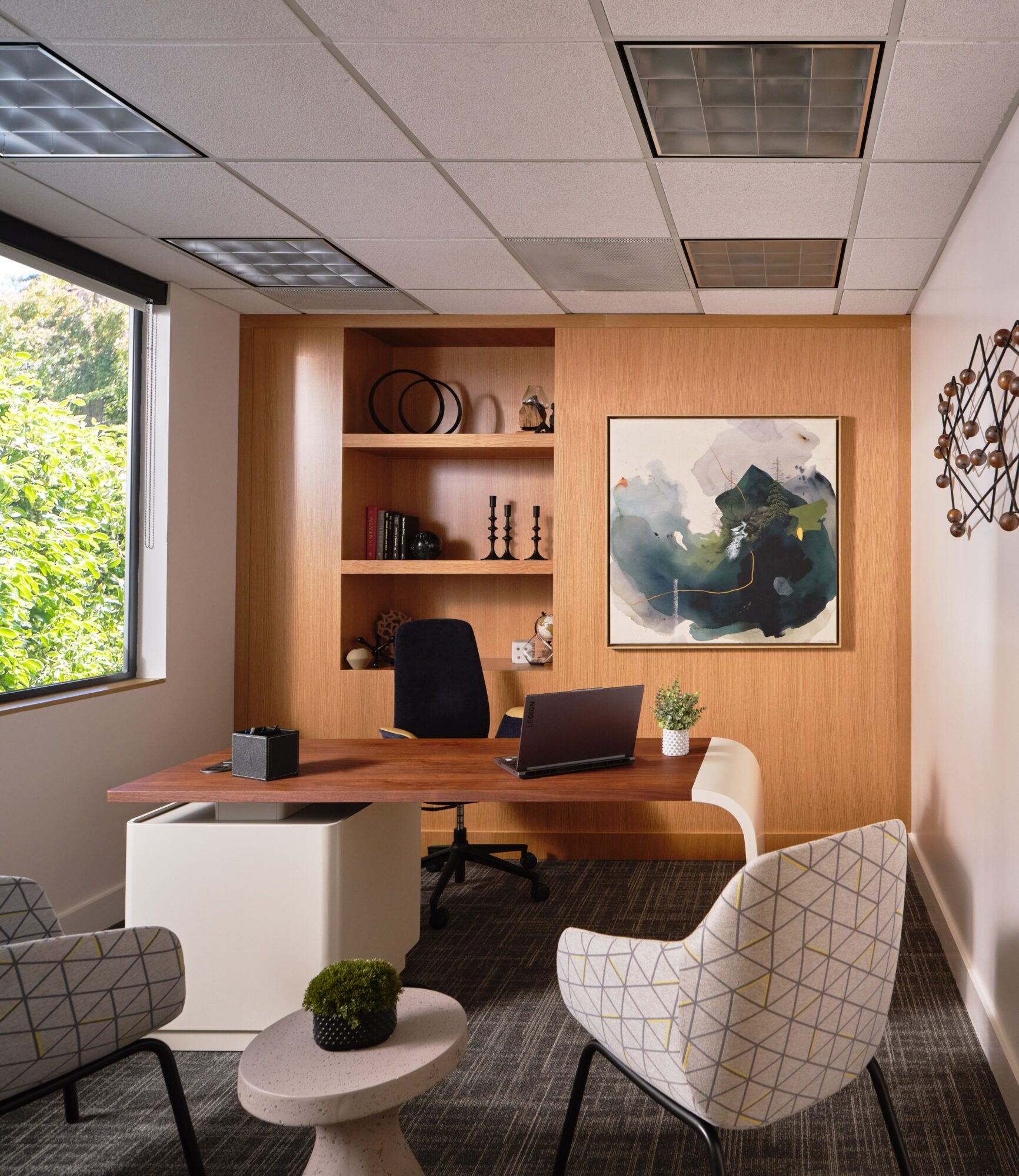 Office with wooden desk, black chair, bookshelf, abstract painting, window with greenery, and two patterned chairs, one with a small plant.