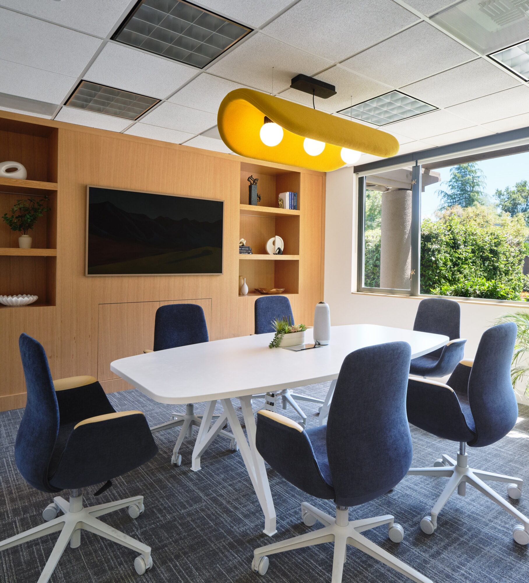 Meeting room with white table, six blue chairs, window view of trees, and yellow ceiling light fixture.