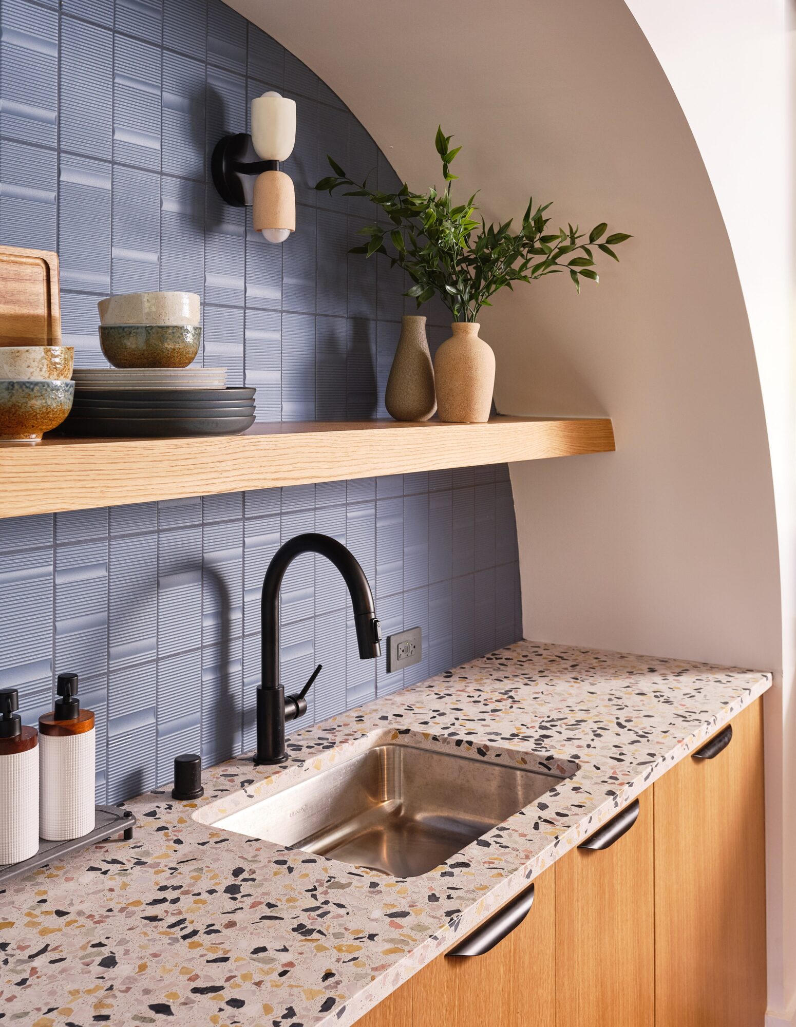 Kitchen countertop with sink, black faucet, potted plant, and shelves with bowls and containers, against a blue tiled wall.