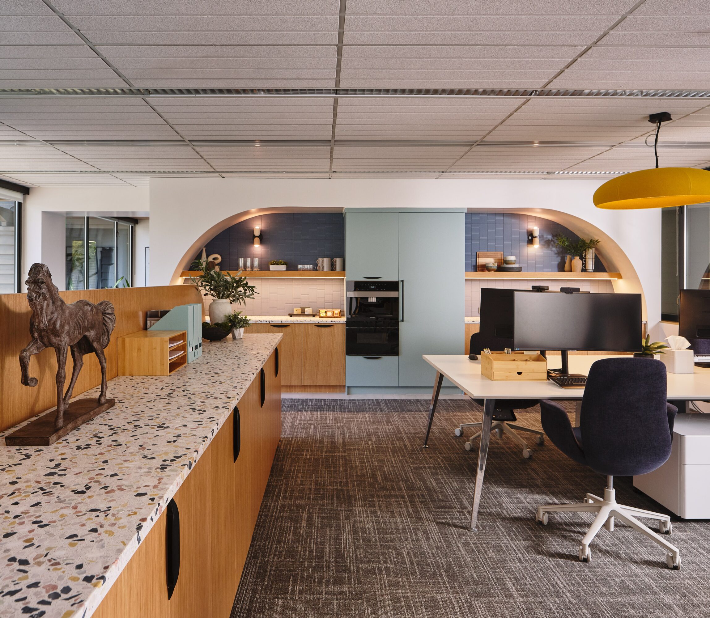 Modern office with a curved wall, desk, computer, and decorative horse sculpture, illuminated by ceiling lights and a yellow pendant lamp.