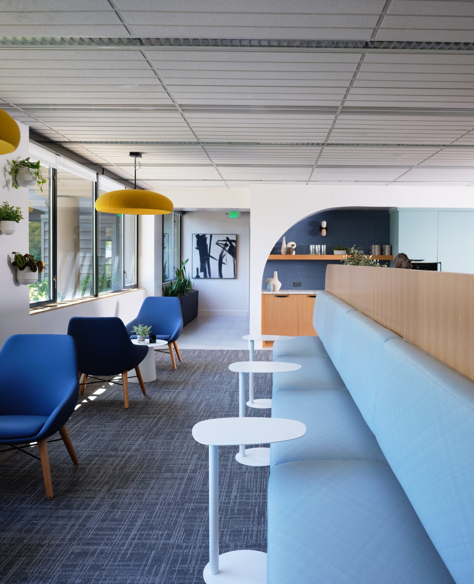 Modern lounge area with blue chairs, yellow pendant lights, and large windows, featuring a reception desk and artwork.