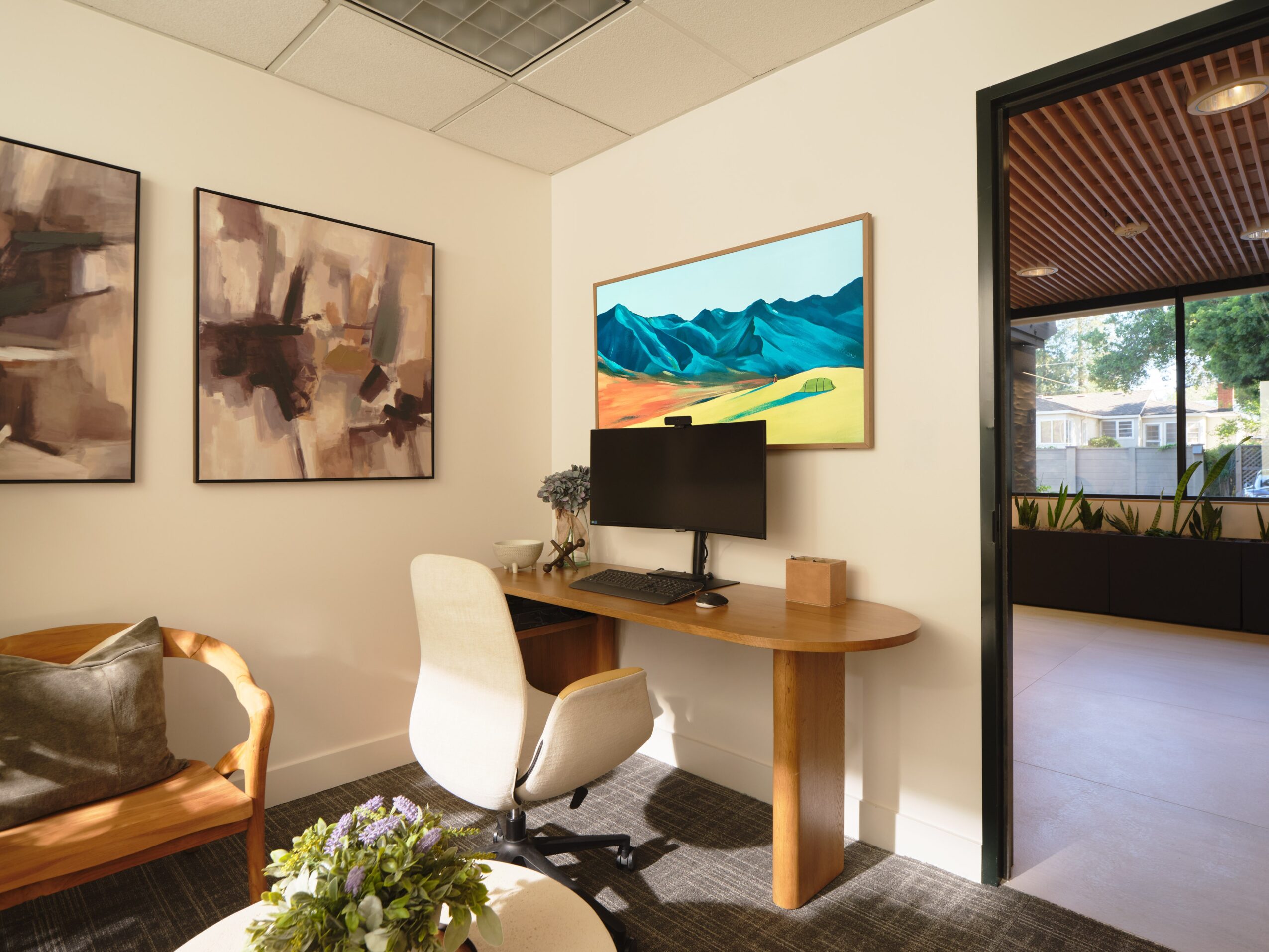 Office corner with paintings, a desk, computer, and a white chair, next to a window with outdoor view.