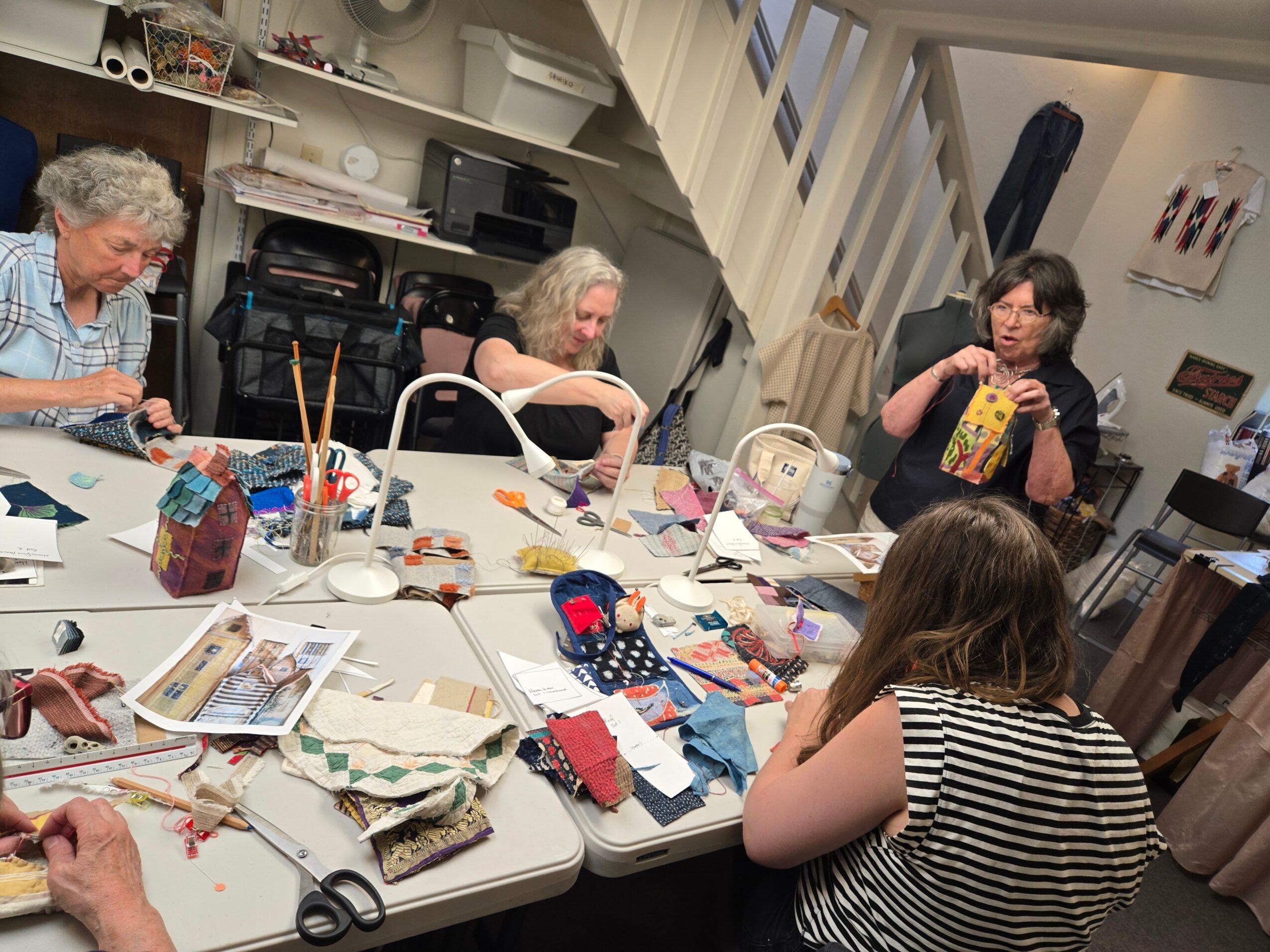 Group of women and a girl sitting around a table with sewing supplies, fabric, and books in a room.