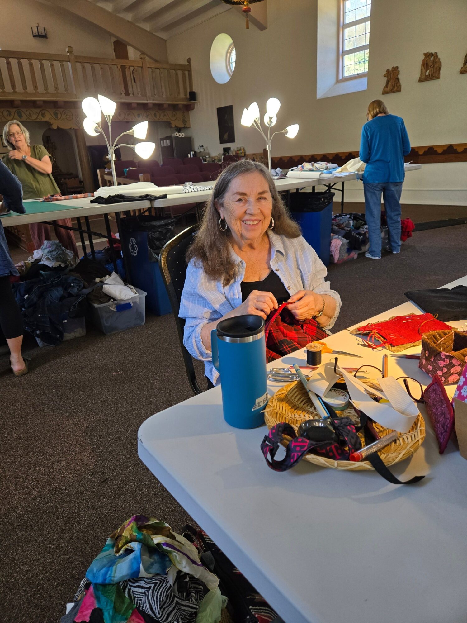 Woman sitting at a table with various colorful fabrics and sewing supplies, in a room with large windows and hanging lights.