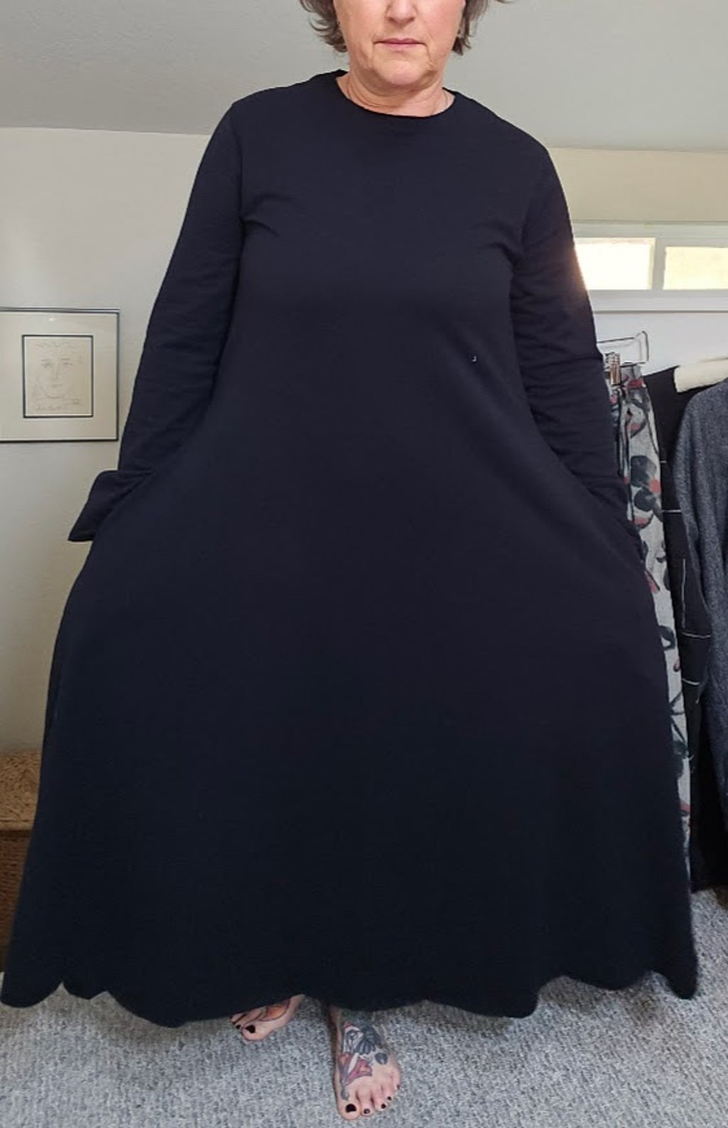 Person standing indoors wearing a long black dress, holding the sides out, with a window and furniture in background.