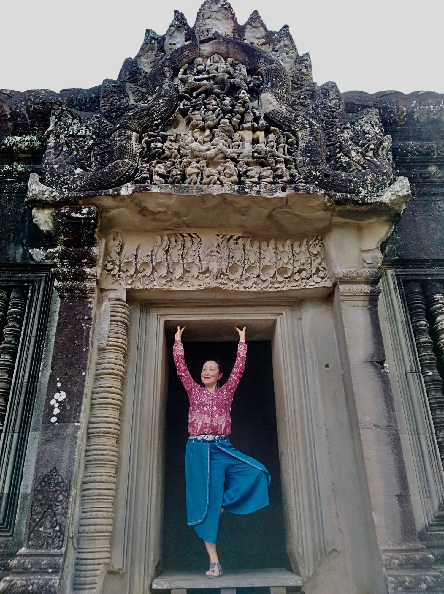 Person standing in a doorway with arms raised, wearing a pink top and blue pants, in front of an ancient stone structure.