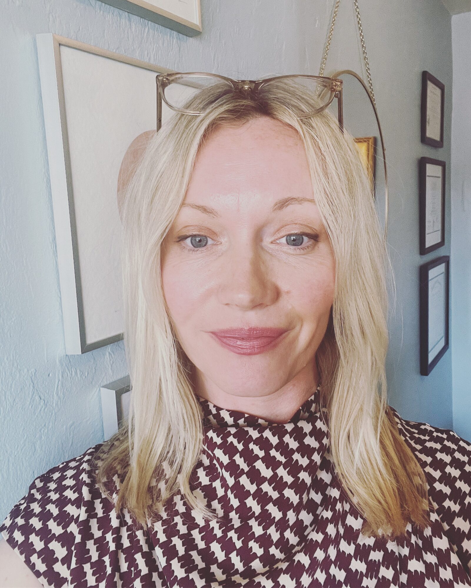 Woman with blonde hair and glasses on head, smiling, wearing a patterned top, in a room with framed pictures on the wall.