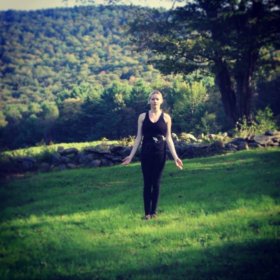 Woman standing on grass with trees and hills in background, facing forward, outdoors during daytime.