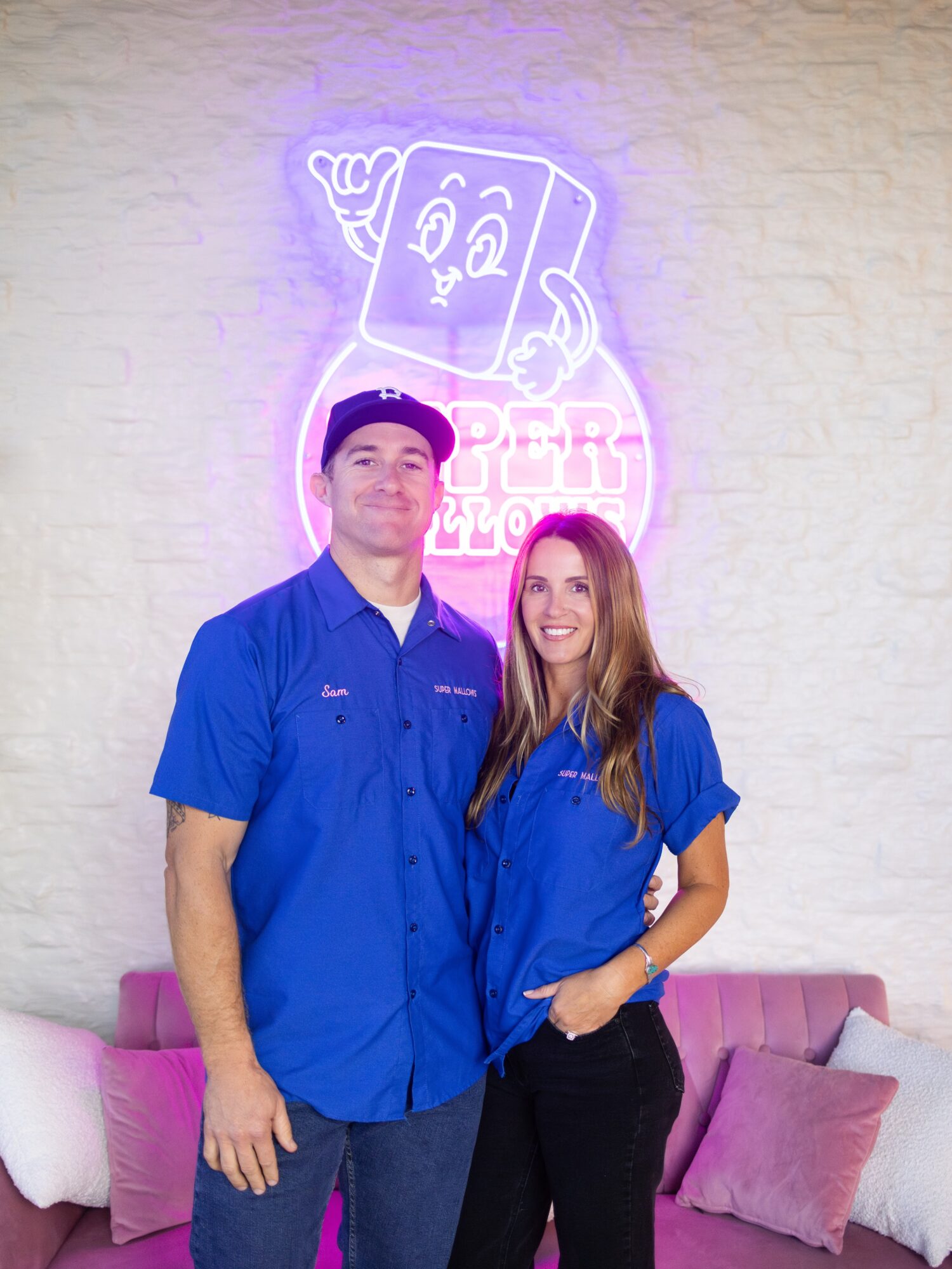 Two people in blue shirts standing in front of a wall with a neon sign of a smiling cartoon character holding a drink, pink and purple lighting.
