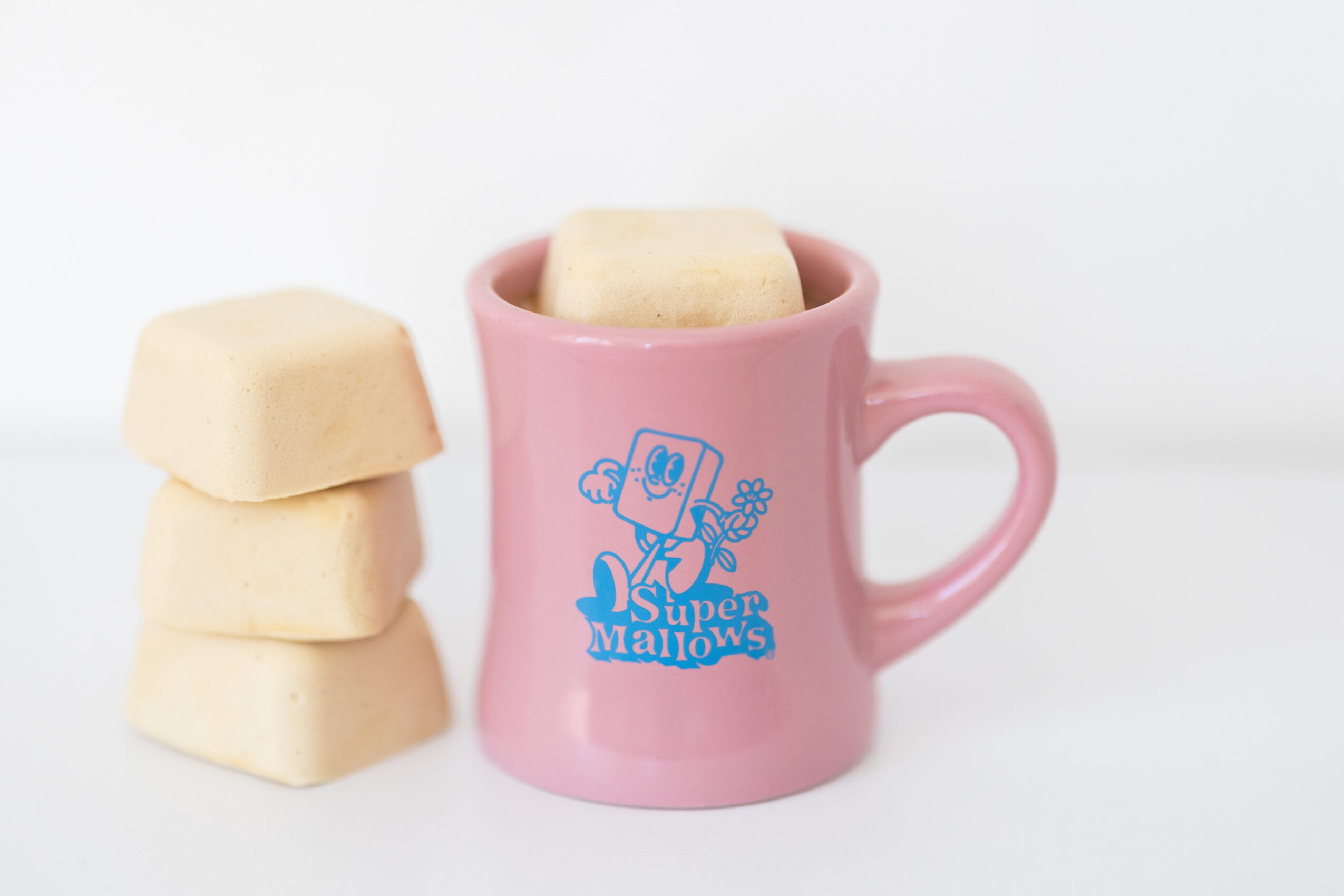 Pink mug with a cartoon character and text, three marshmallows stacked beside it, on a white background.