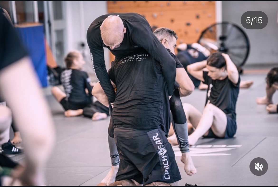 People practicing martial arts or self-defense in a gym, with a focus on a person in black clothing bending forward.
