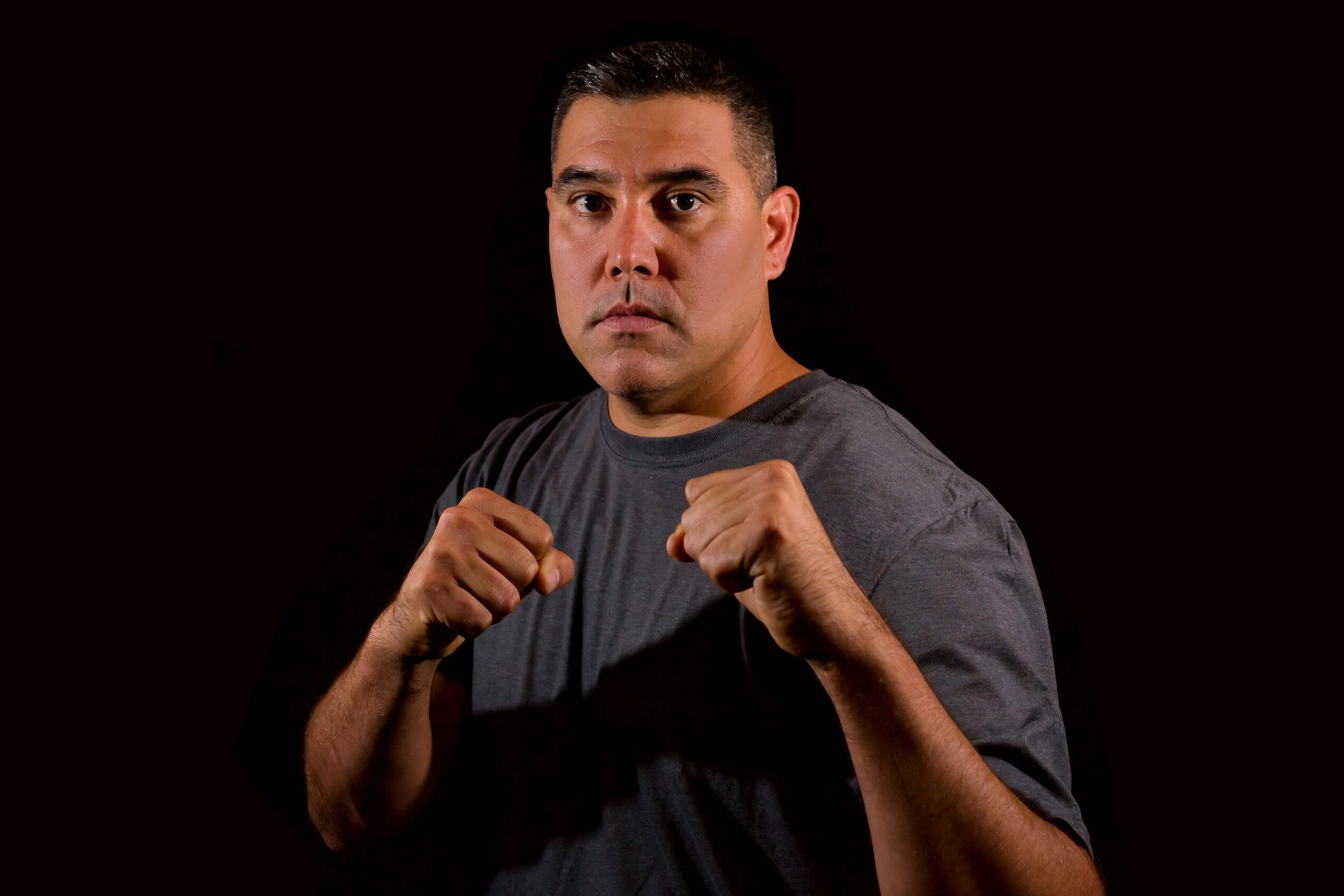 Man in a gray T-shirt with fists raised in a fighting stance, against a black background.