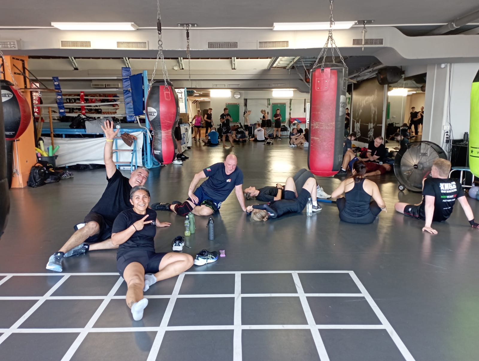 Group of people resting and stretching in a gym with boxing bags and workout equipment, some lying on the floor.