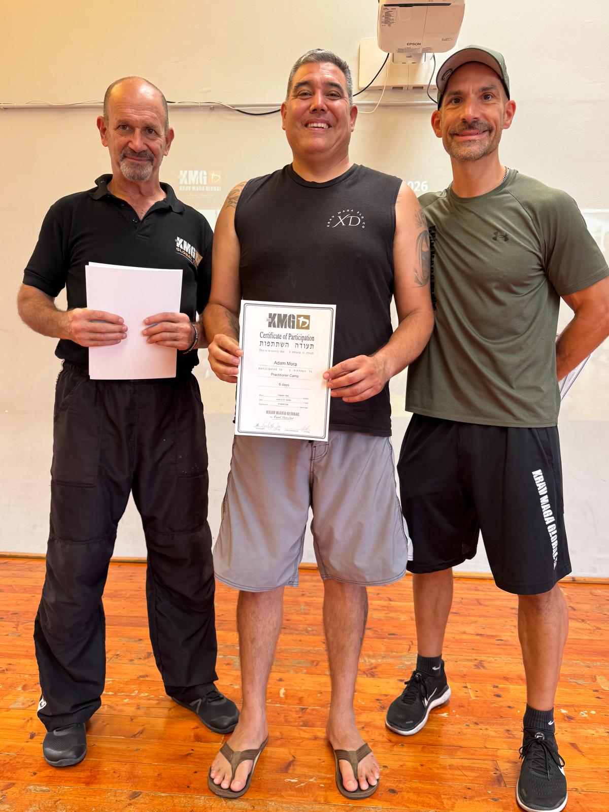 Three men standing indoors, one holding a certificate, smiling, with a wooden floor and a wall with equipment behind them.