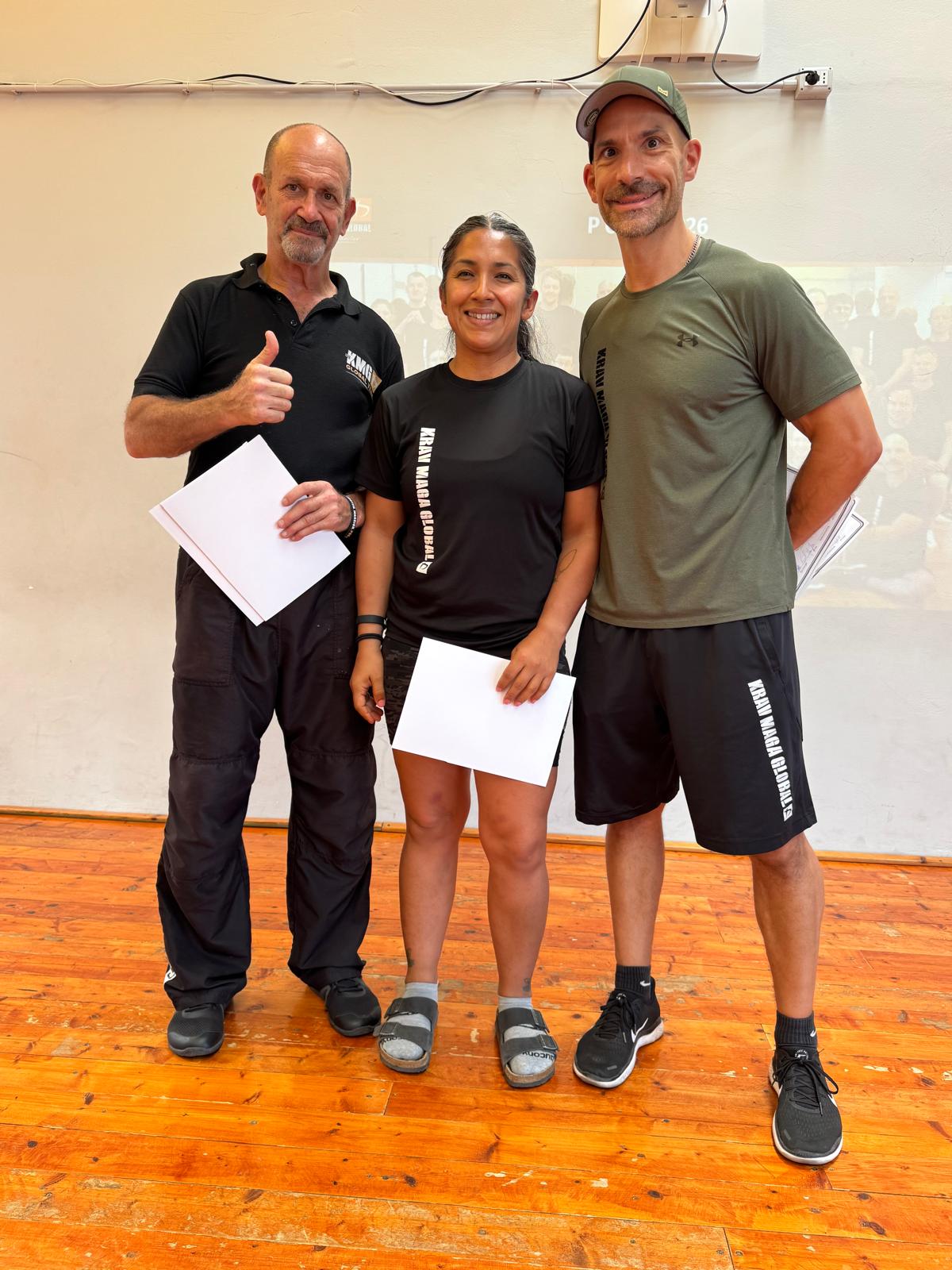 Three people standing indoors on a wooden floor, holding papers, smiling, with a plain wall background.