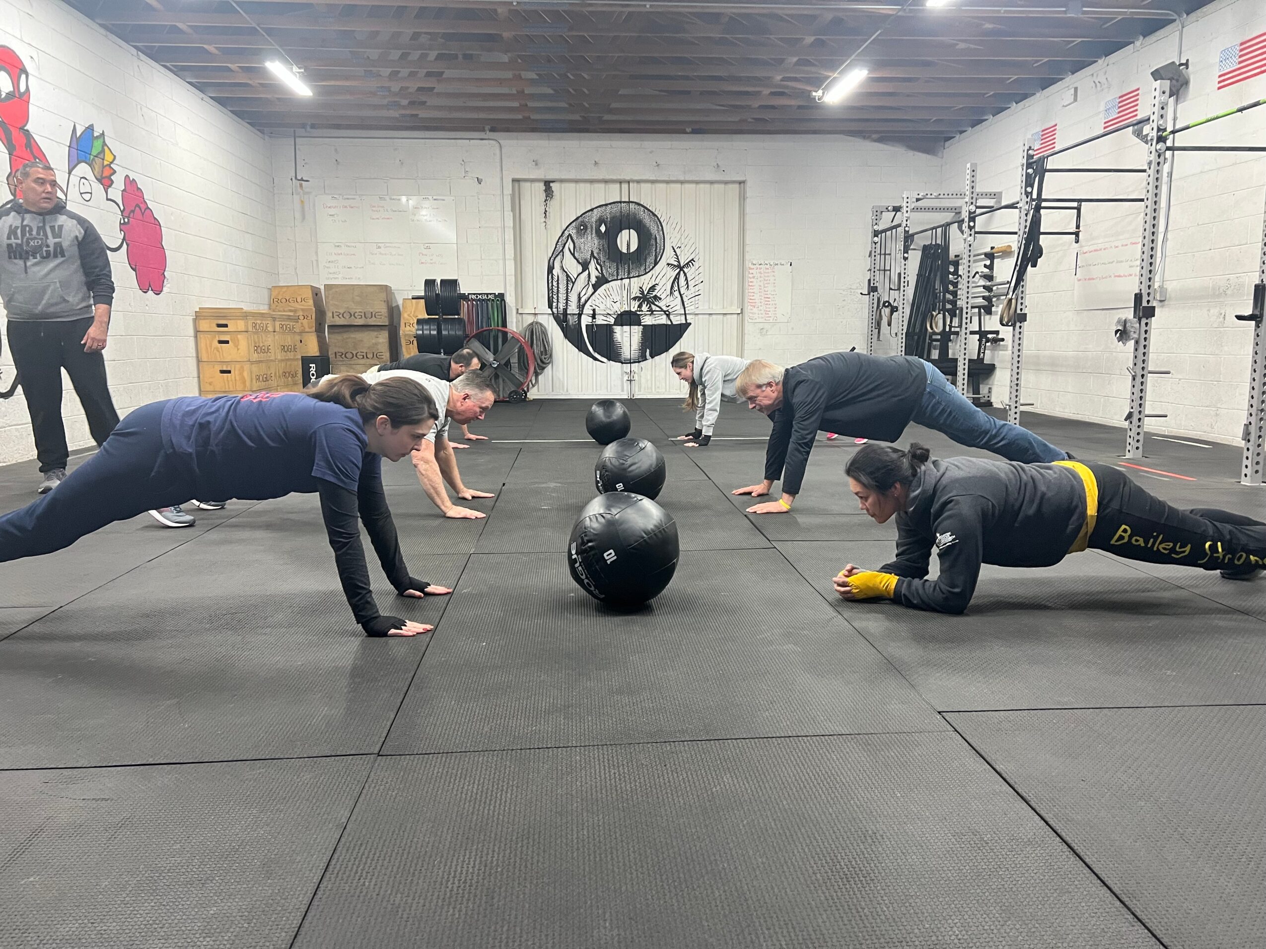 Four people in a gym doing push-ups in a line, with black exercise balls between them, and a person standing in the background.