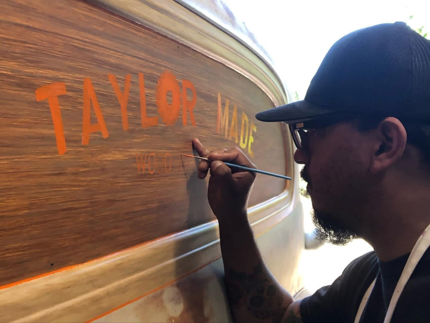 Person wearing a cap and glasses writing on a wooden boat with a paintbrush, spelling 'TAYLOR' in orange letters.