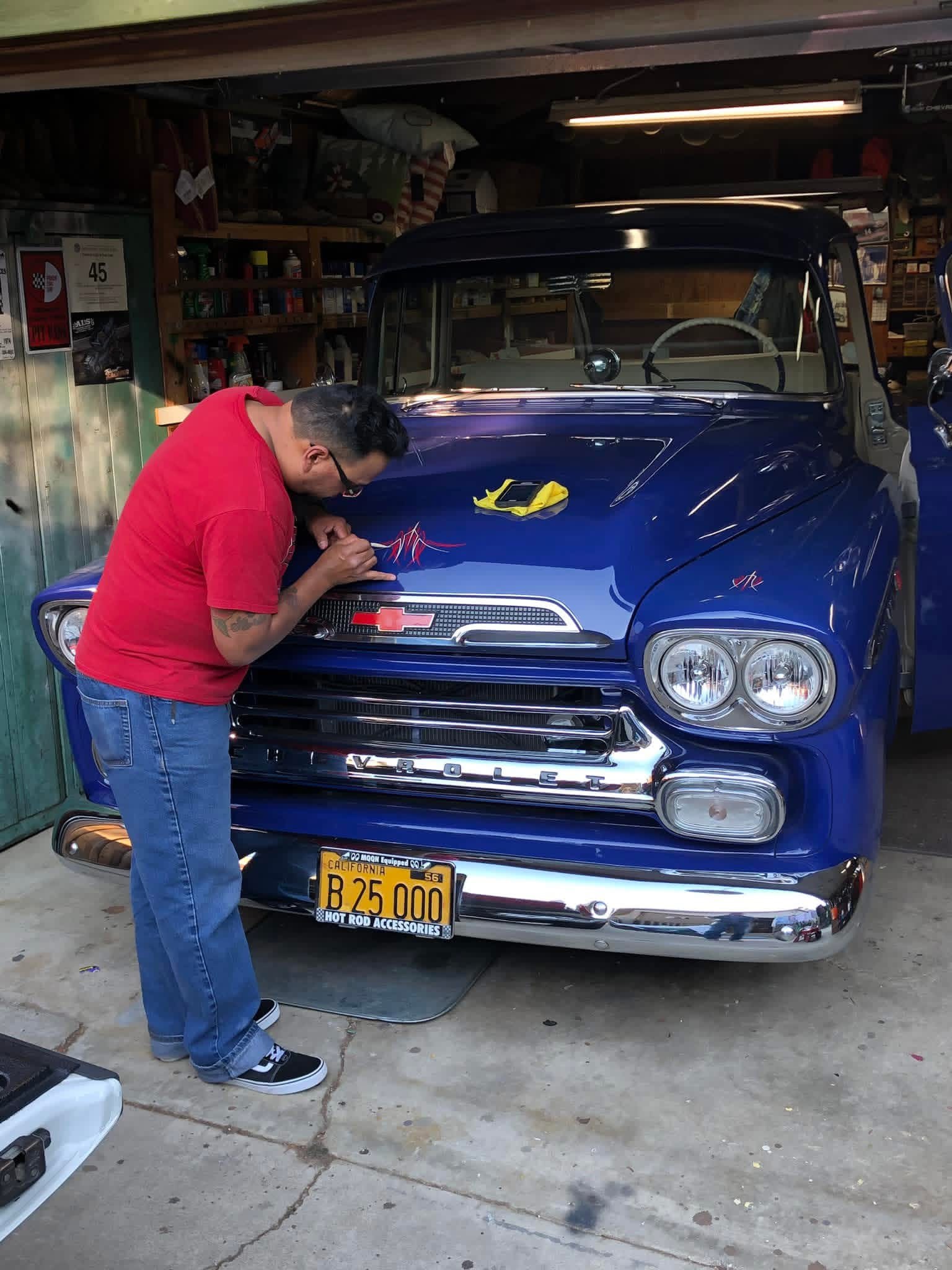 Man in red shirt working on a blue vintage car inside a garage, with shelves and tools in background.