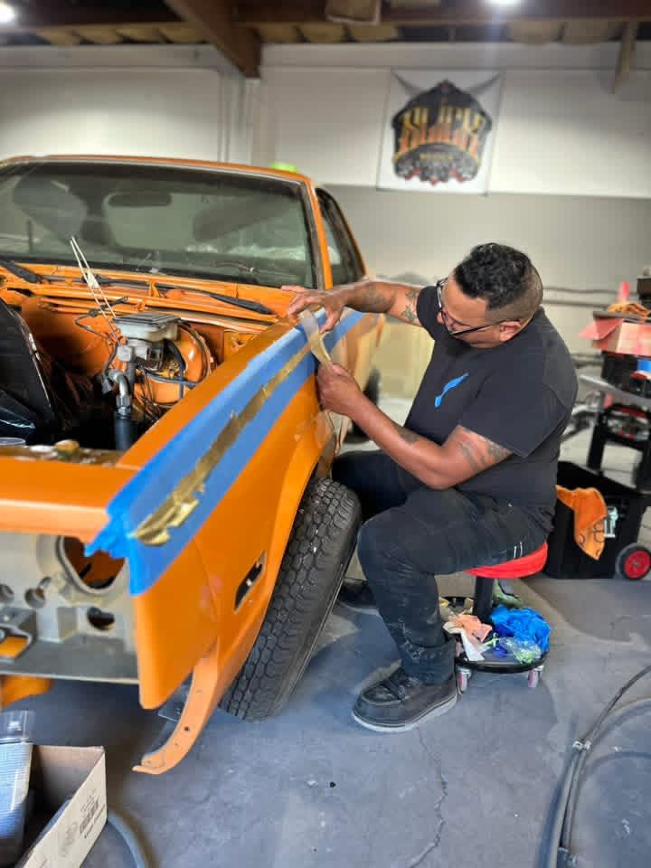 Man working on a car in a garage, using tape on the orange vehicle's side panel, with tools and equipment around.
