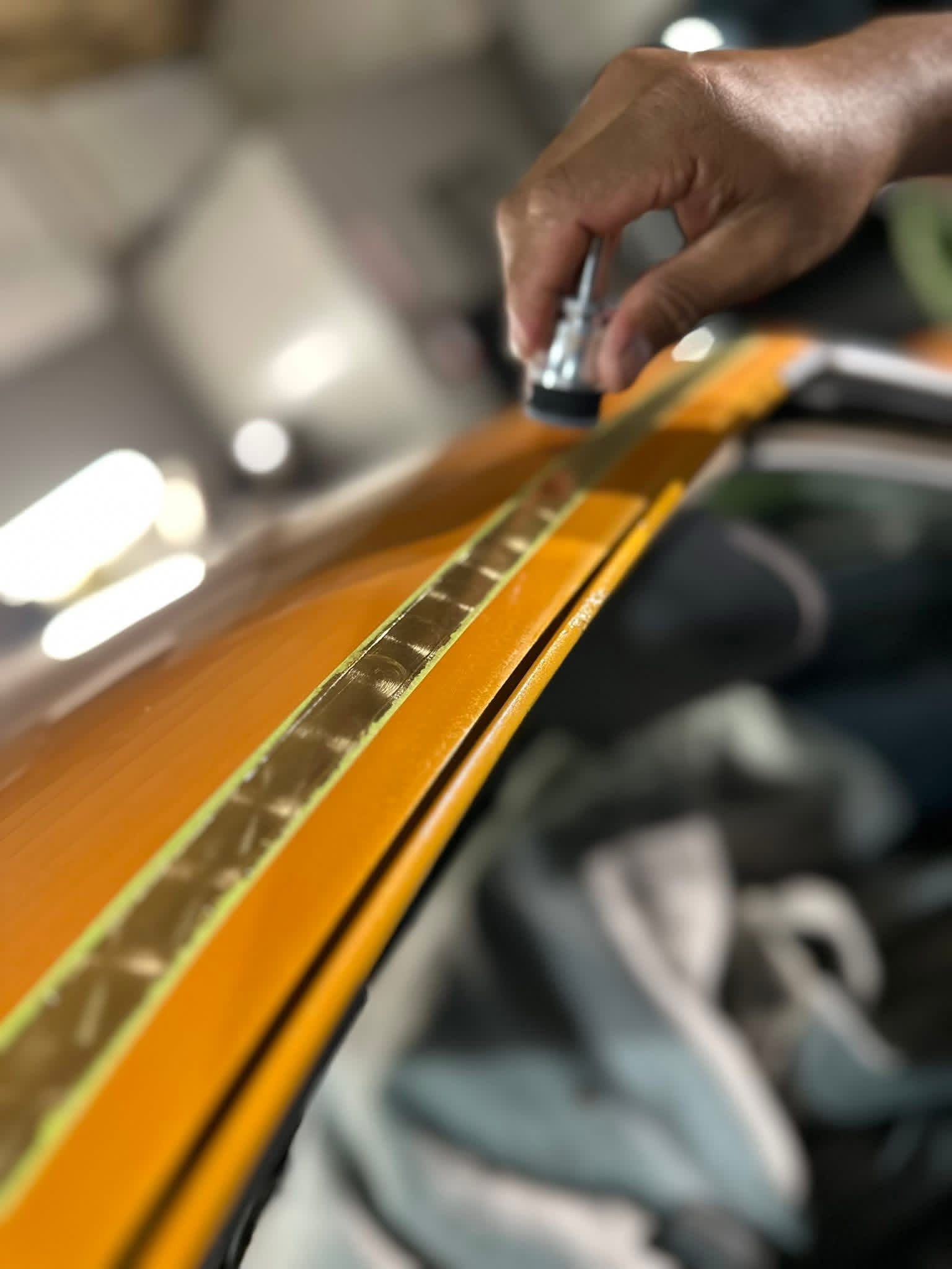 Close-up of a person's hand applying a gold strip to a surface, possibly a vehicle, in a workshop setting.