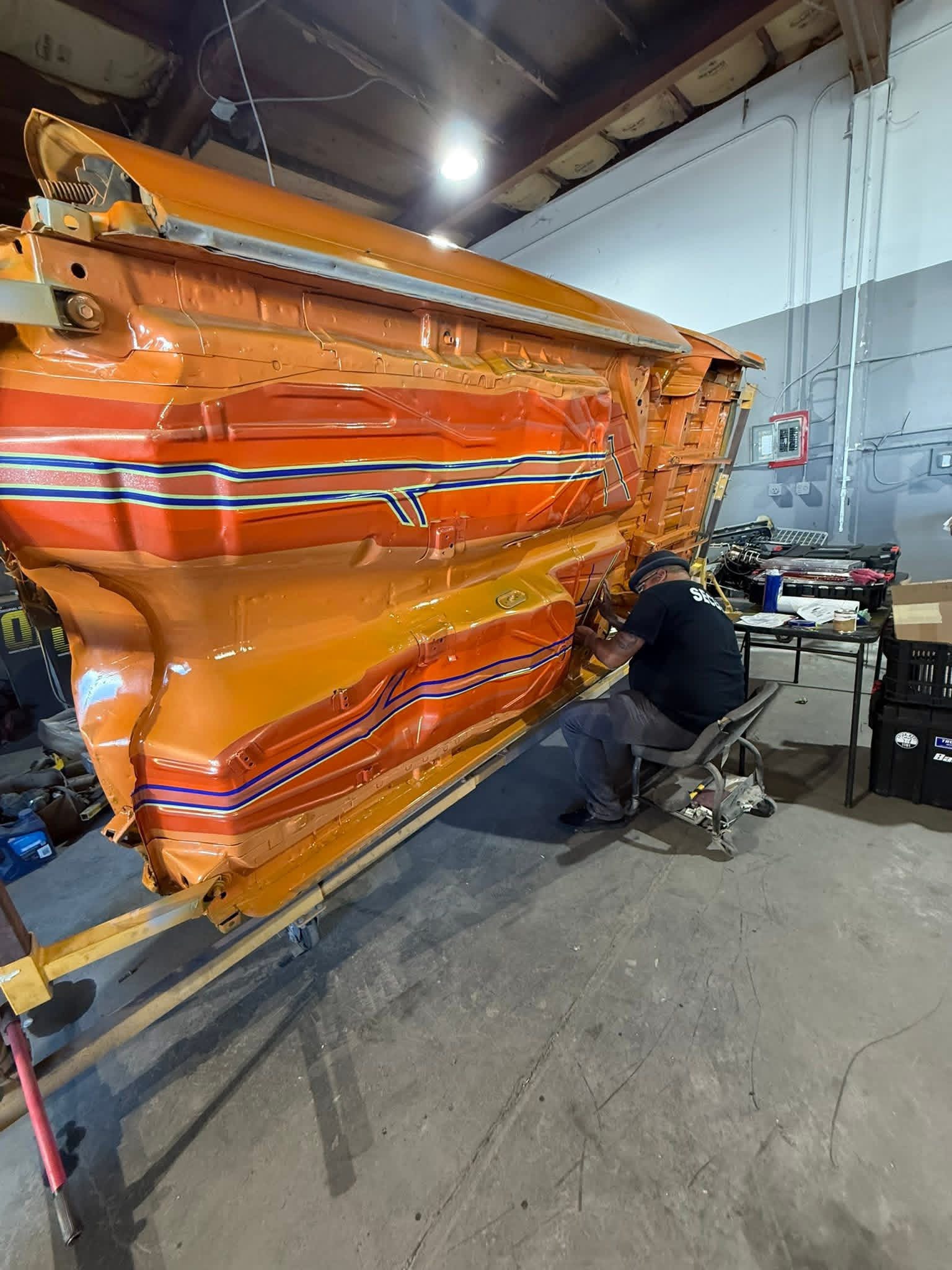 Person working on a large orange and red striped vehicle part inside a workshop, with tools and equipment nearby.
