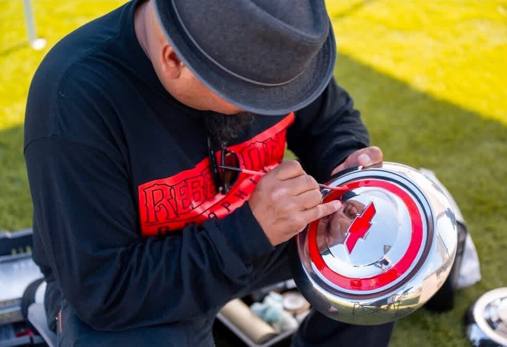 Person wearing a gray hat and black shirt with red text, drawing on a circular object outdoors.